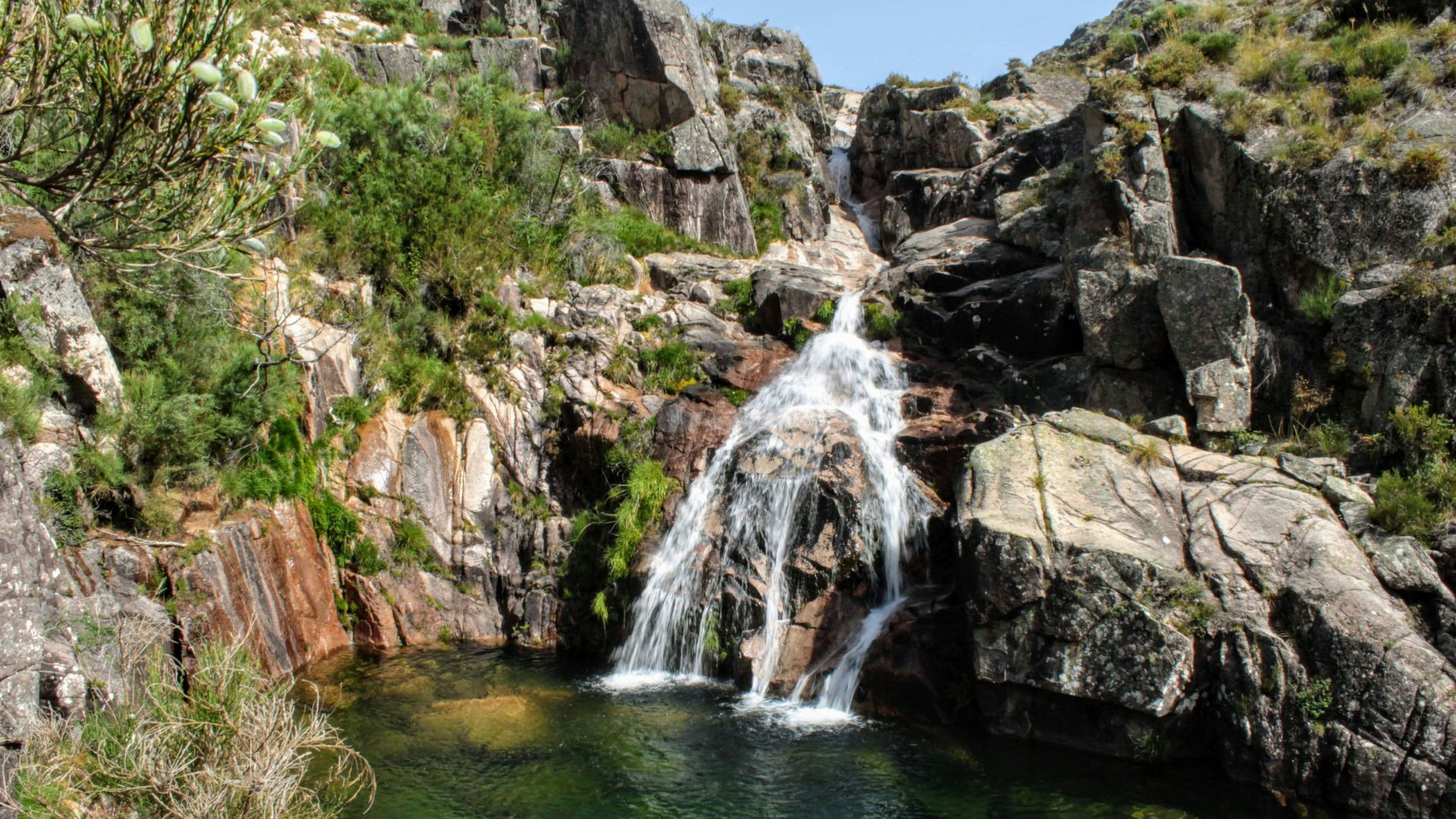 A natural waterfall cascading over rocks into a clear pool in a rocky, green landscape.