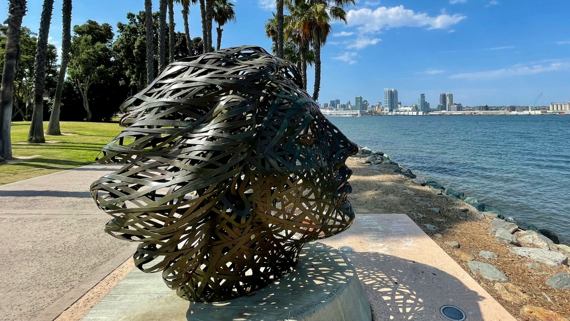 A metal sculpture resembling a human head made of interwoven bands, set against a backdrop of a bay with a city skyline and palm trees under a blue sky.