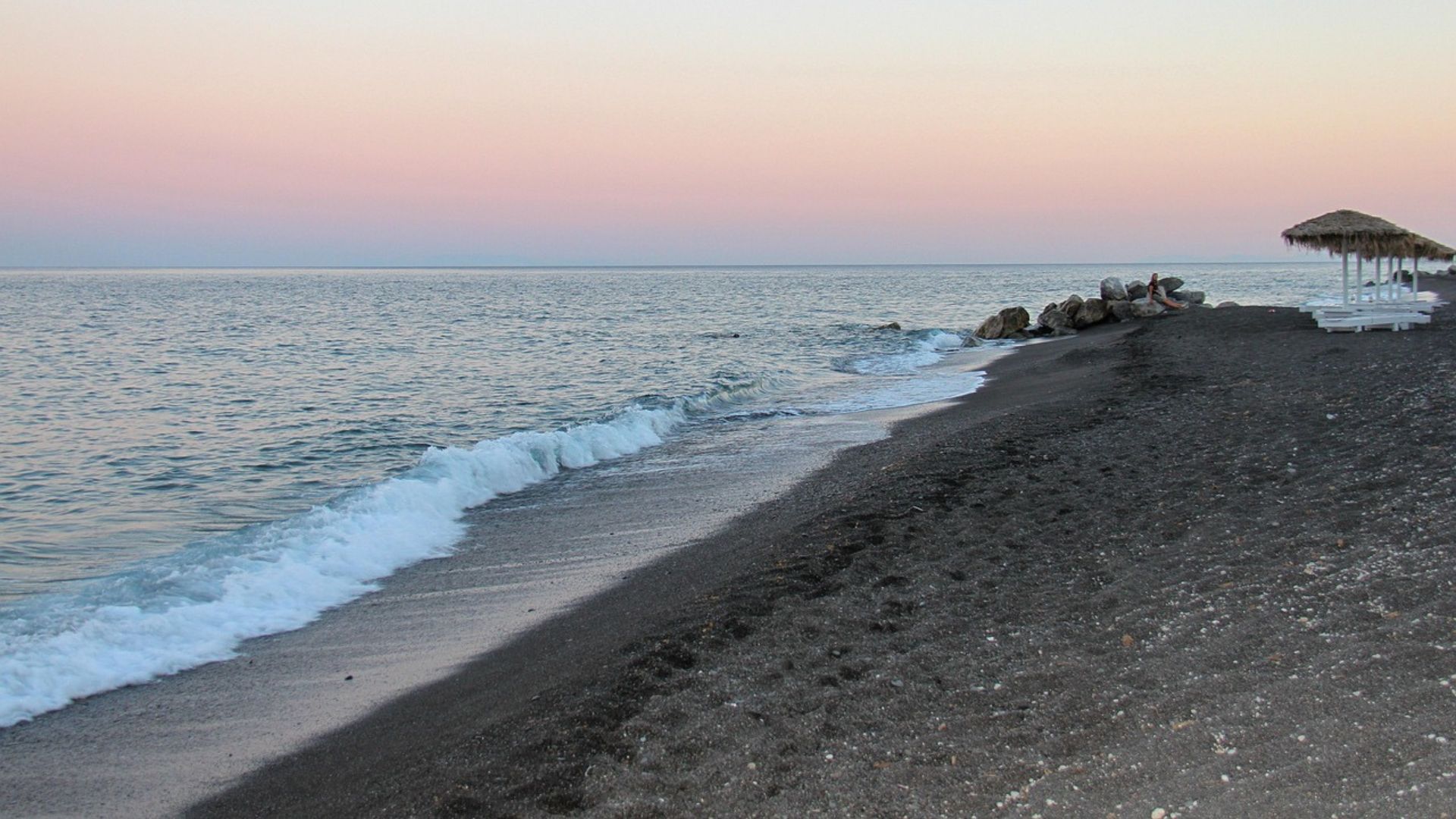 A serene view of Perissa Beach at dusk, with black sand and a calm sea.