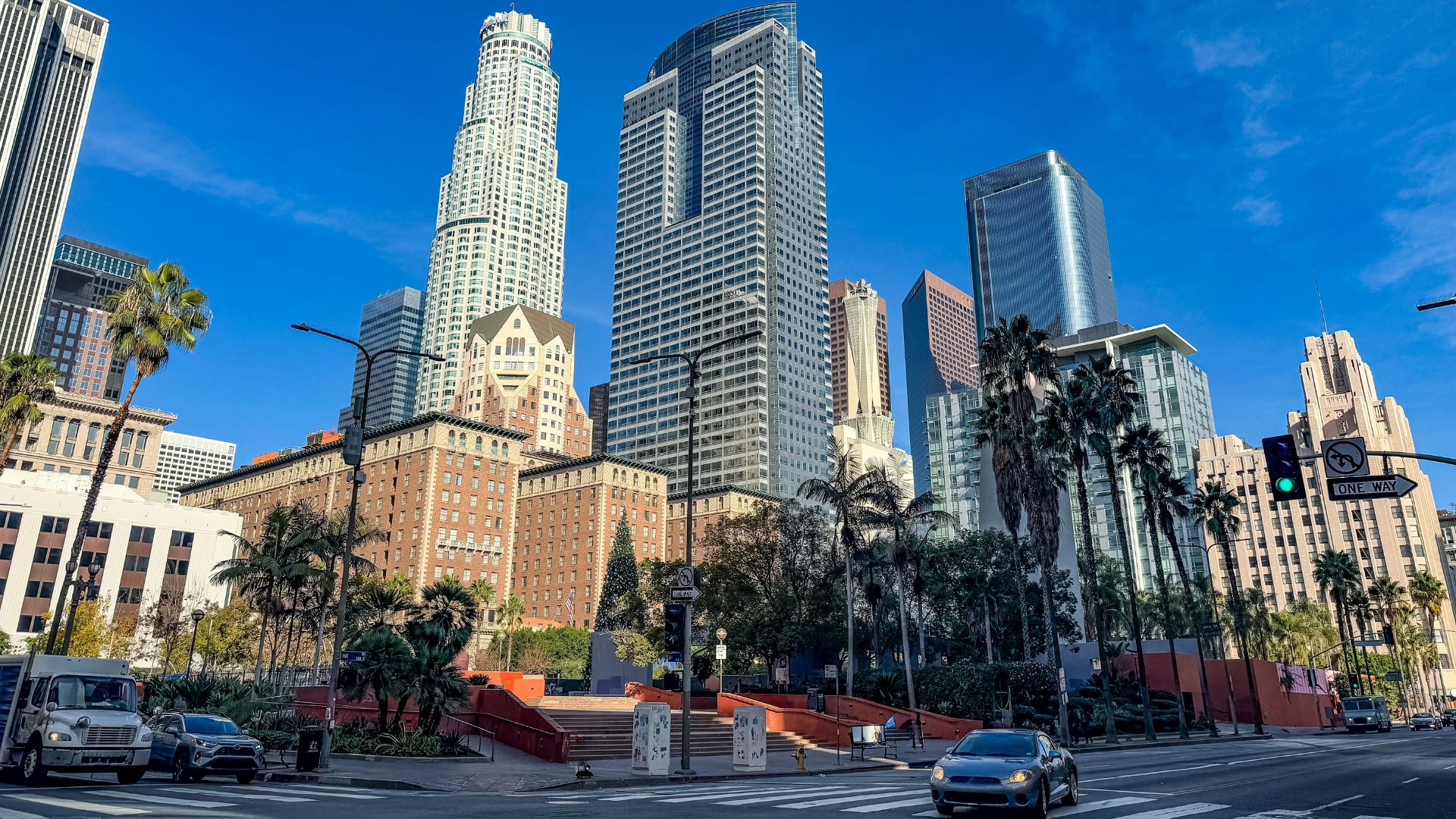 A sunny wide shot of Pershing Square in Downtown Los Angeles, showing the park area with palm trees, street, and several prominent skyscrapers including the U.S. Bank Tower and Gas Company Tower under a clear blue sky. Cars are visible on the street in the foreground.