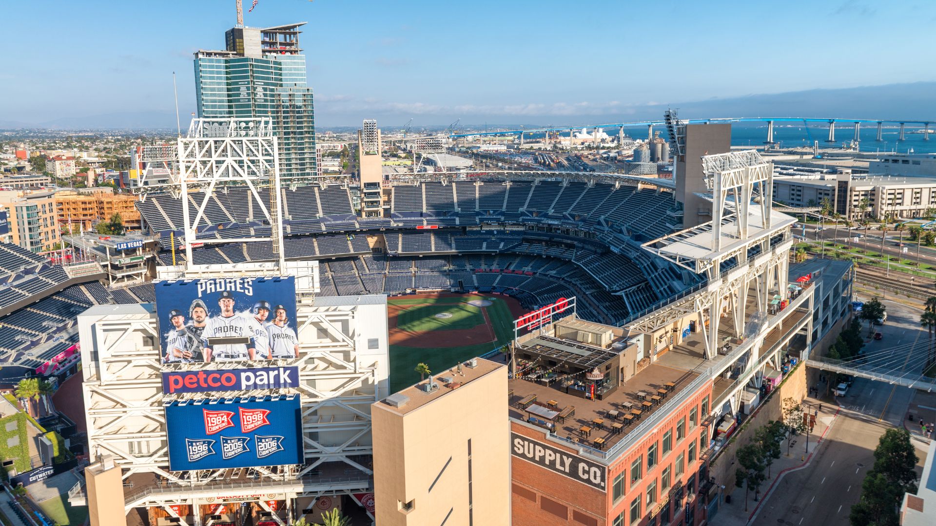 An aerial view of Petco Park, home of the San Diego Padres, nestled within the downtown San Diego skyline, with the bay and Coronado Bridge visible in the background.