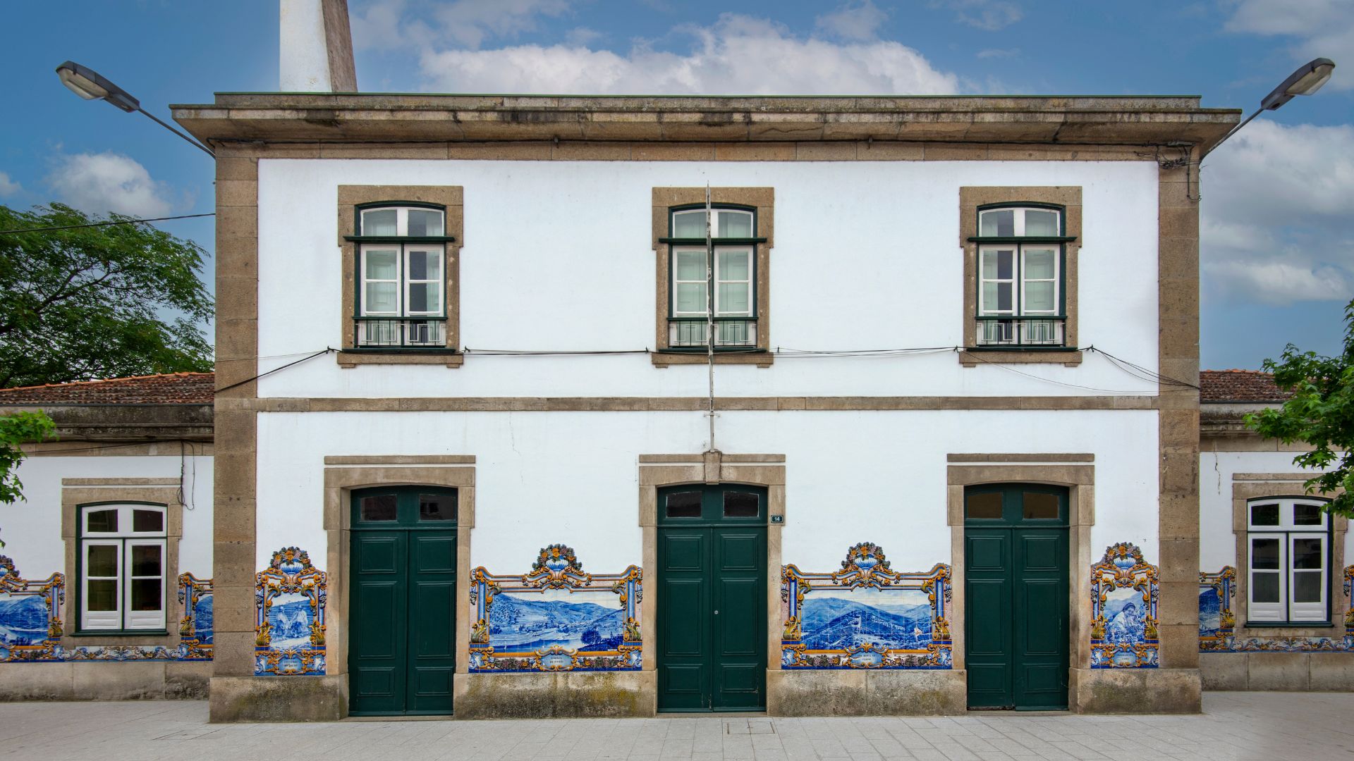 A white building with green doors and windows, decorated with blue and white azulejo tiles depicting scenes of the Douro Valley, under a partly cloudy sky.