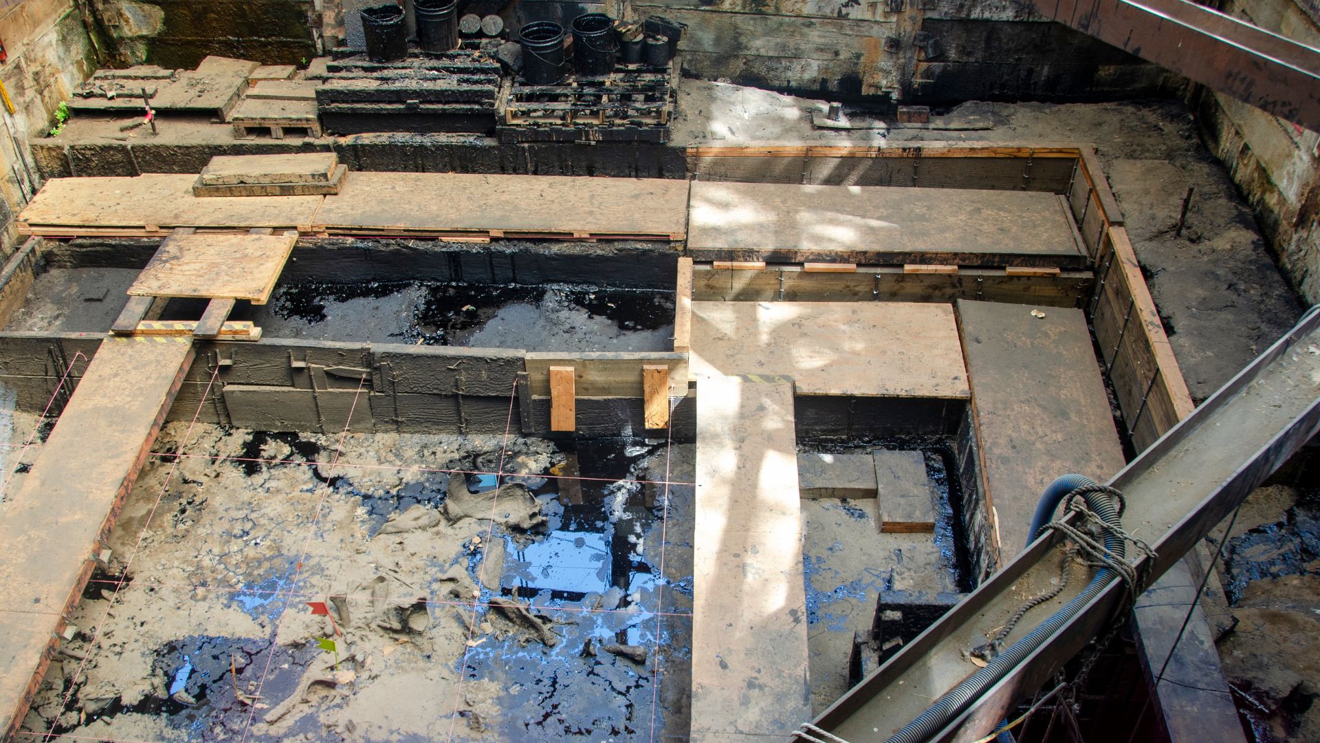 Aerial view of Pit 91 at the La Brea Tar Pits in Los Angeles, showing an active archaeological excavation site with visible tar pits, wooden platforms for access, and exposed fossil finds, suitable for educational or scientific content related to paleontology and natural history.