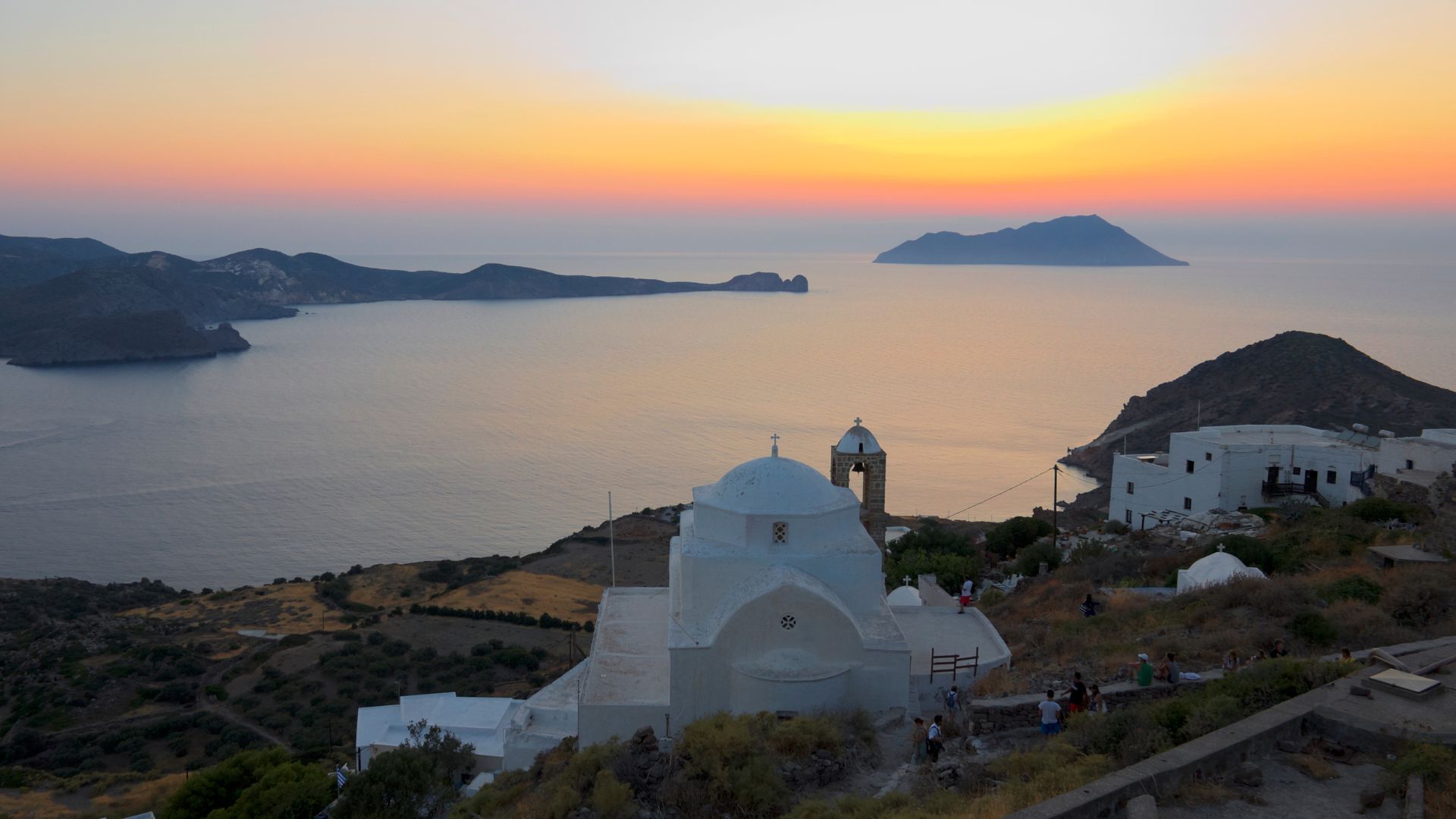 A picturesque white church with a brick bell tower sits atop a hill overlooking the Aegean Sea in Milos, Greece.