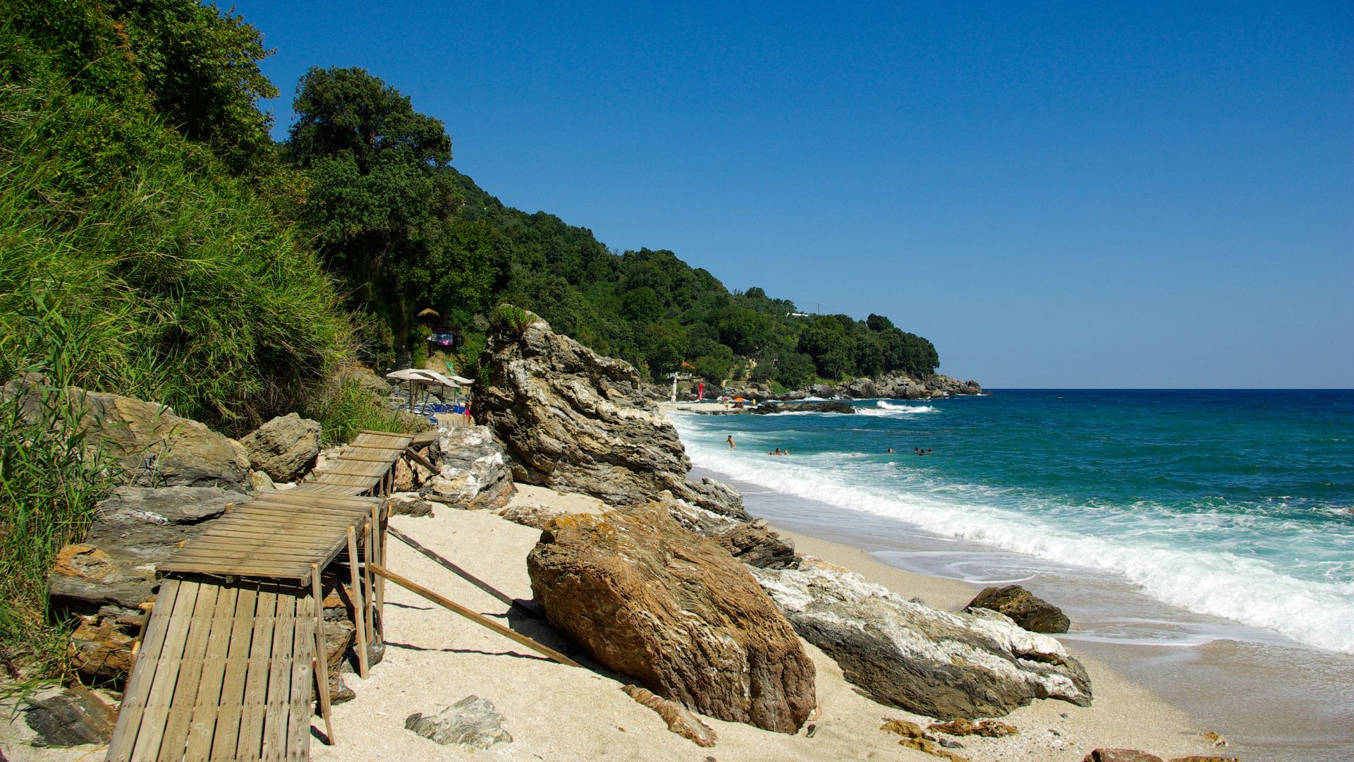 A scenic view of a sandy beach with clear blue waters, rocky outcrops, and a wooden boardwalk leading to the shore.