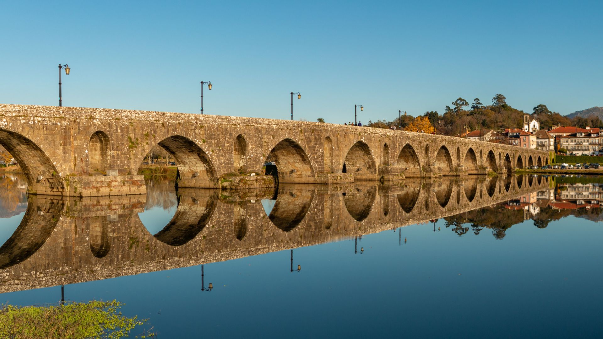 A long, ancient stone bridge with numerous arches spans a calm river, reflecting the bridge and the clear blue sky above. The bridge is illuminated by streetlights, and a town with buildings and trees is visible on the far bank under a bright sky.