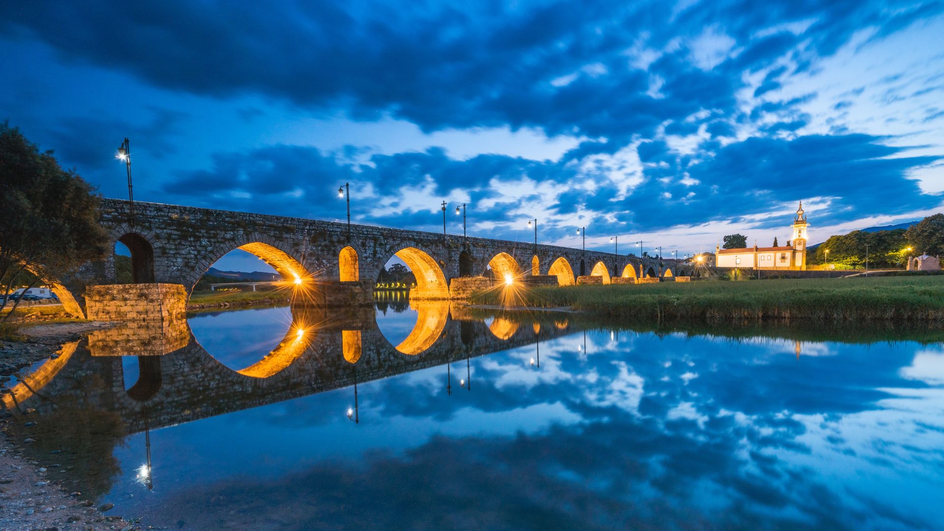 A long stone bridge with multiple arches, illuminated by warm lights, reflects in the calm water of a river under a dramatic blue evening sky. In the background, a small church tower is visible on the riverbank.