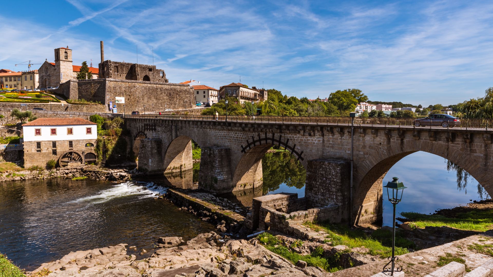 A wide shot of the medieval Ponte de Barcelos (Barcelos Bridge) crossing the Cávado River in Portugal, with a historic town and castle ruins visible on the far bank.