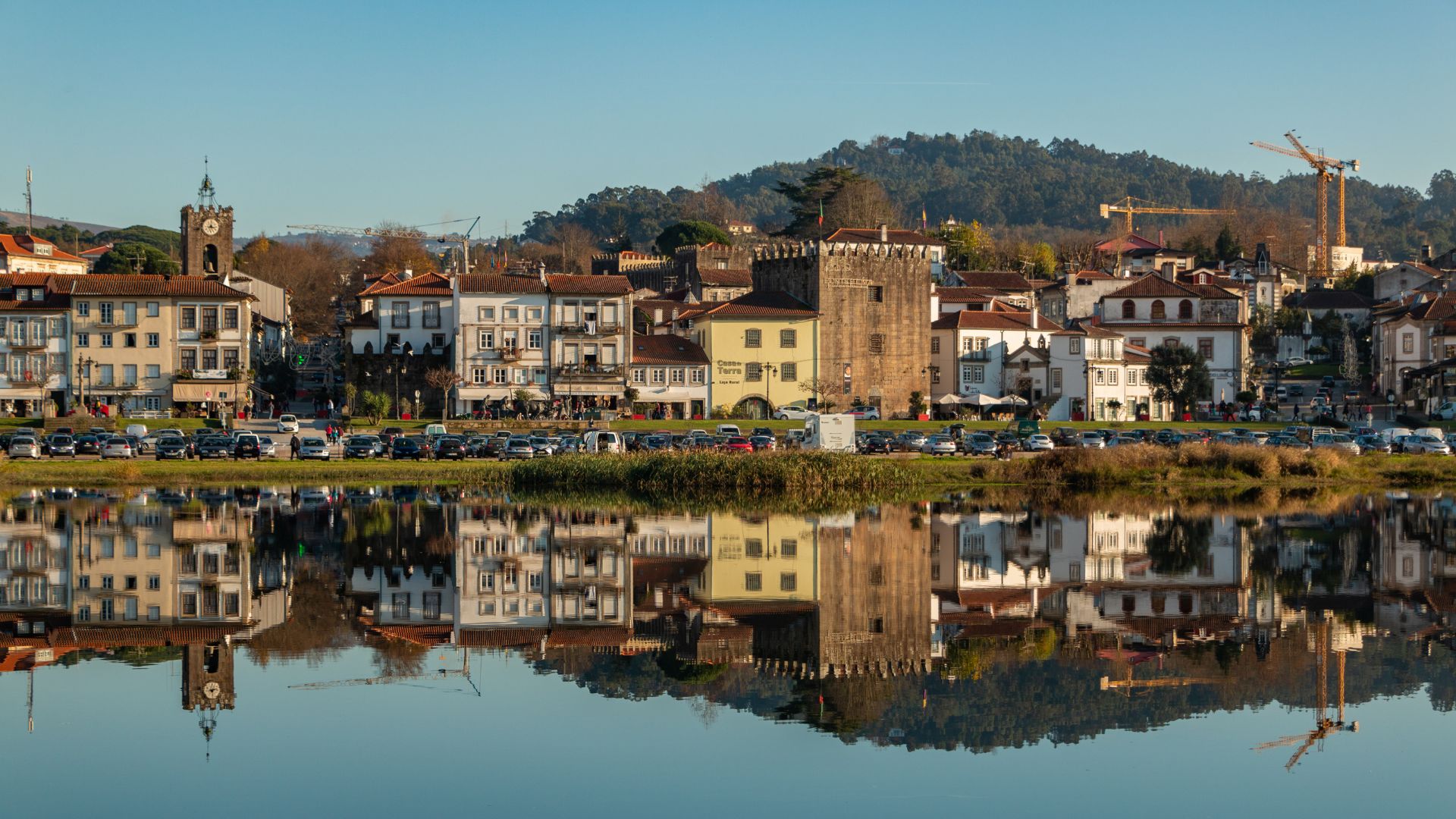 A picturesque view of Ponte de Lima, Portugal, featuring historic buildings and a prominent clock tower reflected in the calm waters of the Lima River, with a lush, green hillside in the background.