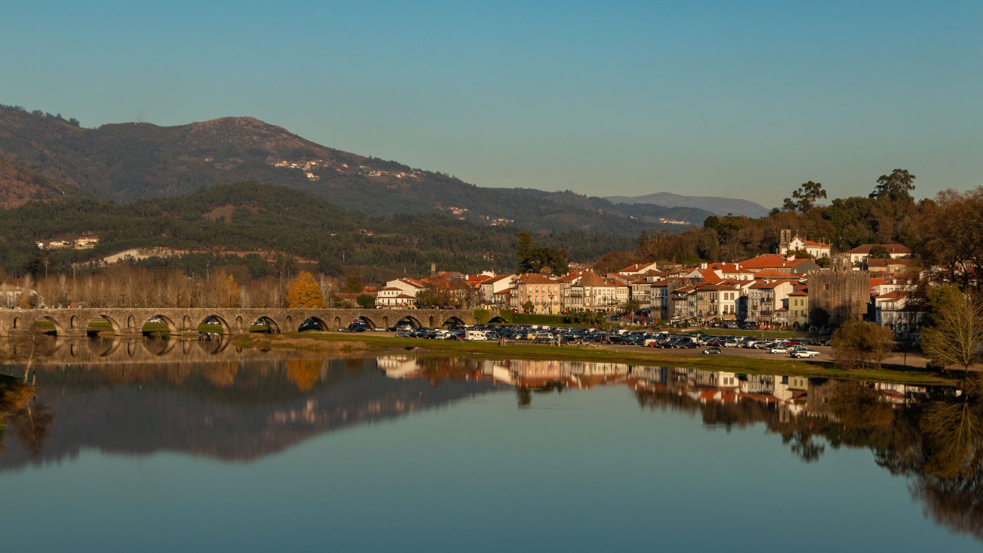 A picturesque view of the historic Roman and Medieval bridge over the Lima River in Ponte de Lima, Portugal, with the town's buildings and distant mountains reflected in the water under a clear sky.