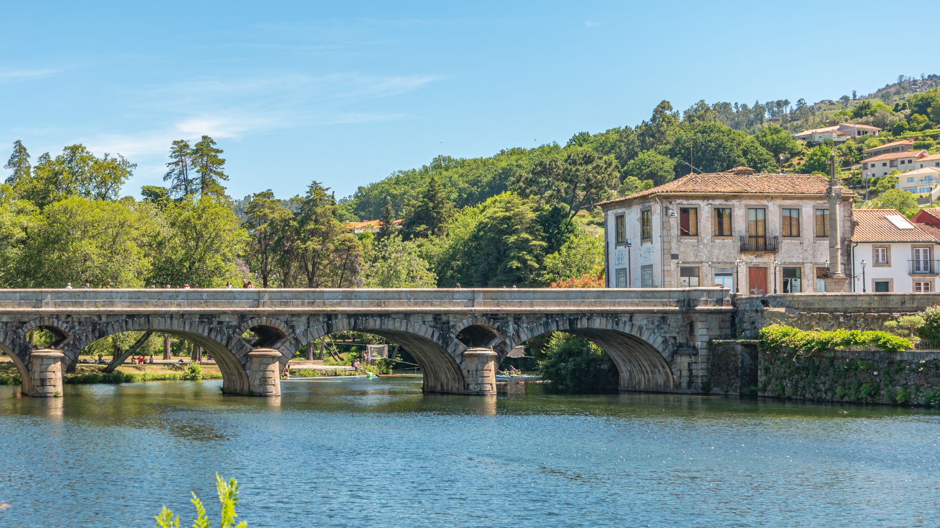 A stone bridge with multiple arches spans a river, with a historic building on the bank and green hills in the background.