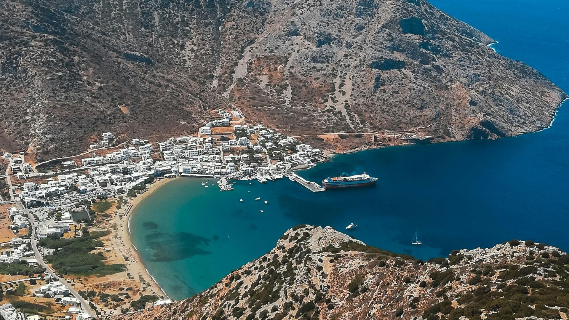 Aerial view of Kamares, a picturesque village on Sifnos island, Greece, featuring a sandy beach, harbor, and surrounding hills.