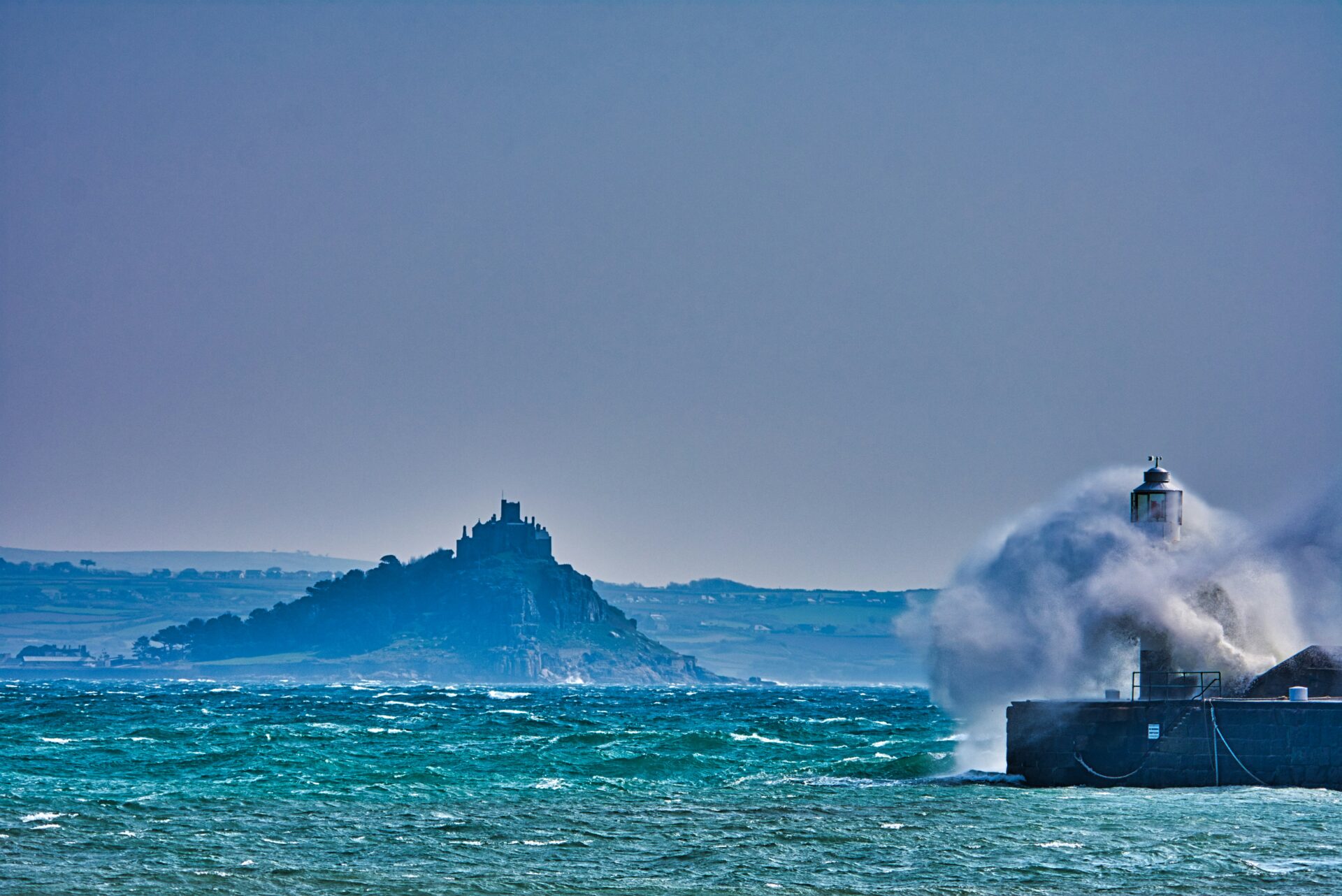 Wave crashes against lighthouse at Penzance, with St Michael’s Mount in the background.