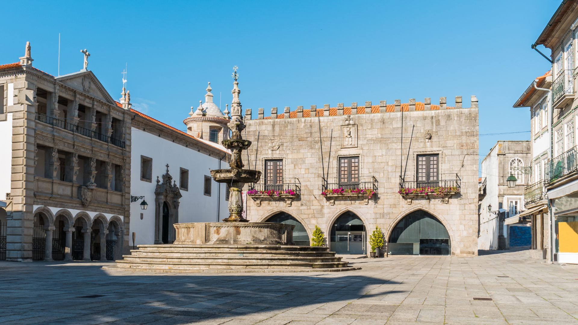 A historic town square in Viana do Castelo, Portugal, featuring an ornate fountain and traditional stone buildings under a clear sky.