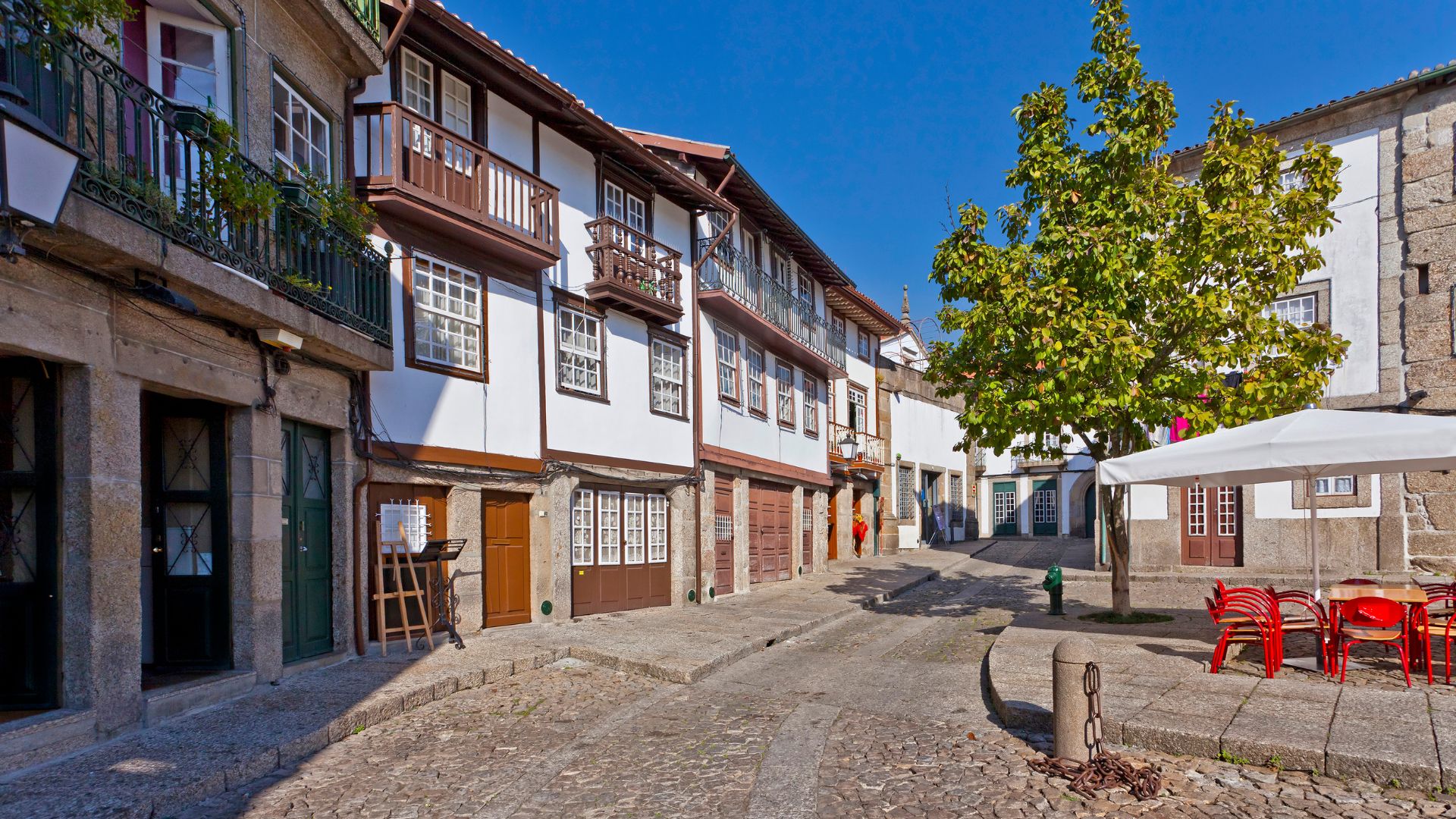 A cobblestone square in Guimarães, Portugal, featuring historic white buildings with balconies and a tree with red outdoor seating under a white umbrella.