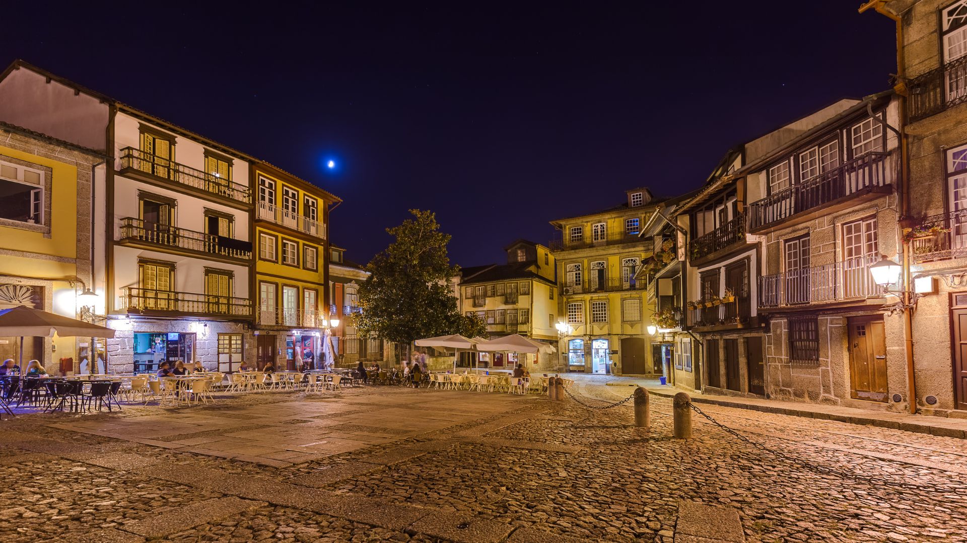Night view of Praça de São Tiago in Guimarães, Portugal, featuring illuminated historic buildings and cobblestone ground under a moonlit sky.