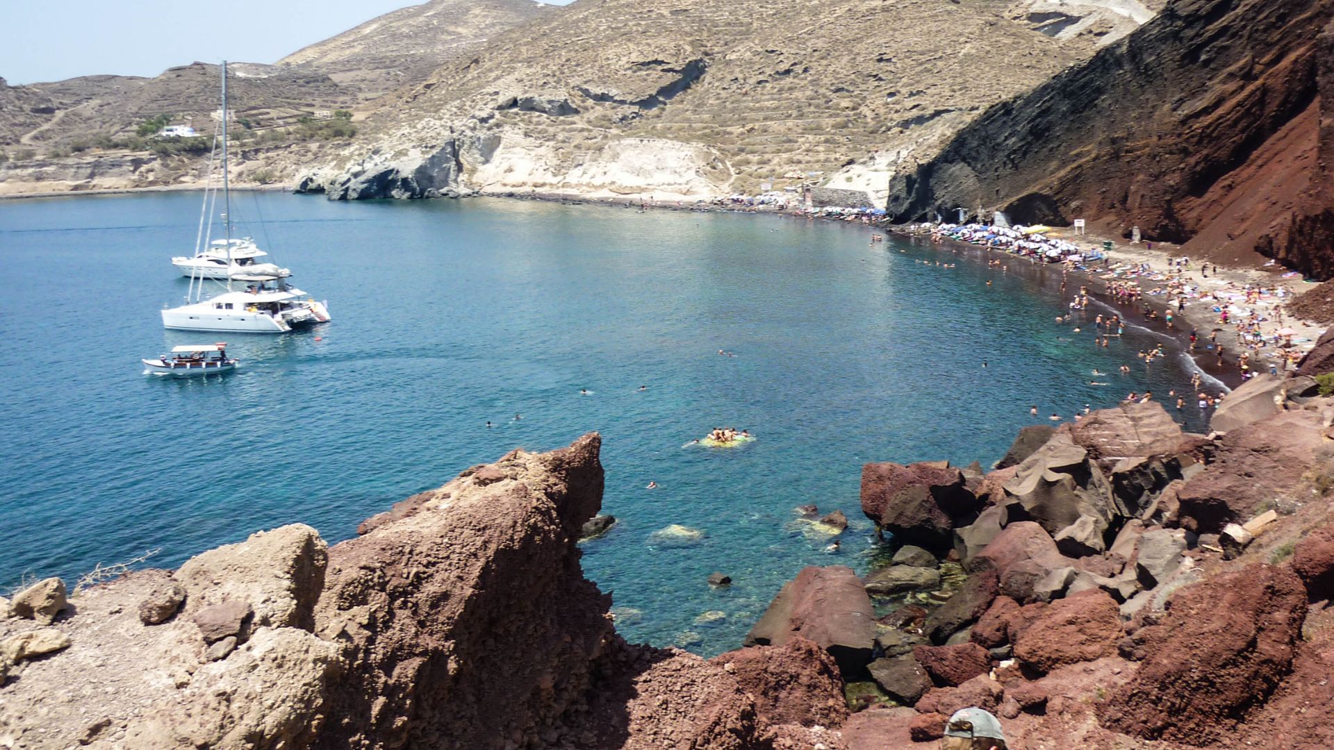 The rocky Red Beach in Santorini, Greece with red cliffs, blue water, boats, and many beach goers.