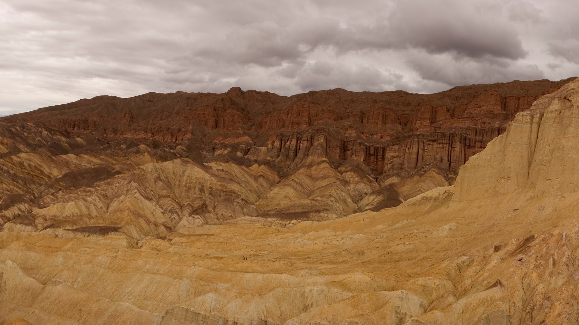 A wide-angle landscape view of Golden Canyon in Death Valley National Park, featuring the towering, reddish-brown rock formations of the Red Cathedral under a dramatic, cloudy sky. The foreground shows a vast expanse of light-colored, arid badlands with intricate erosion patterns.