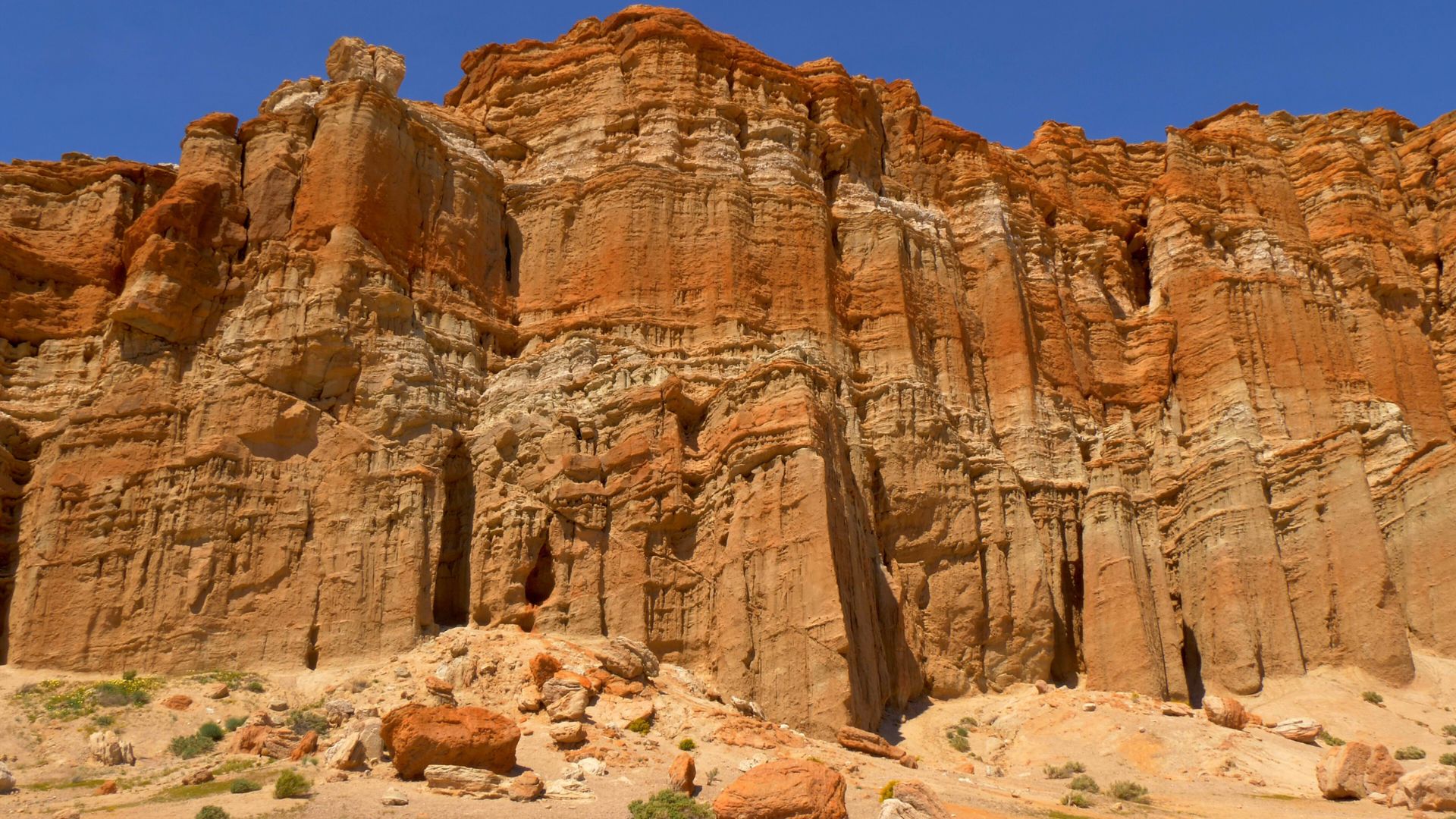 A wide shot of towering, layered red and white rock formations under a clear blue sky, with sparse desert vegetation and scattered rocks in the foreground.