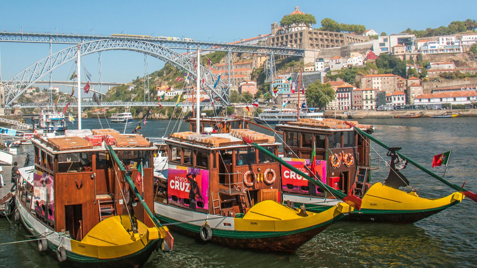 A vibrant scene of traditional rabelo boats on the Douro River in Porto, Portugal, with the iconic Dom Luís I Bridge and colorful cityscape in the background. These boats, historically used for transporting port wine, are a popular tourist attraction for river cruises.