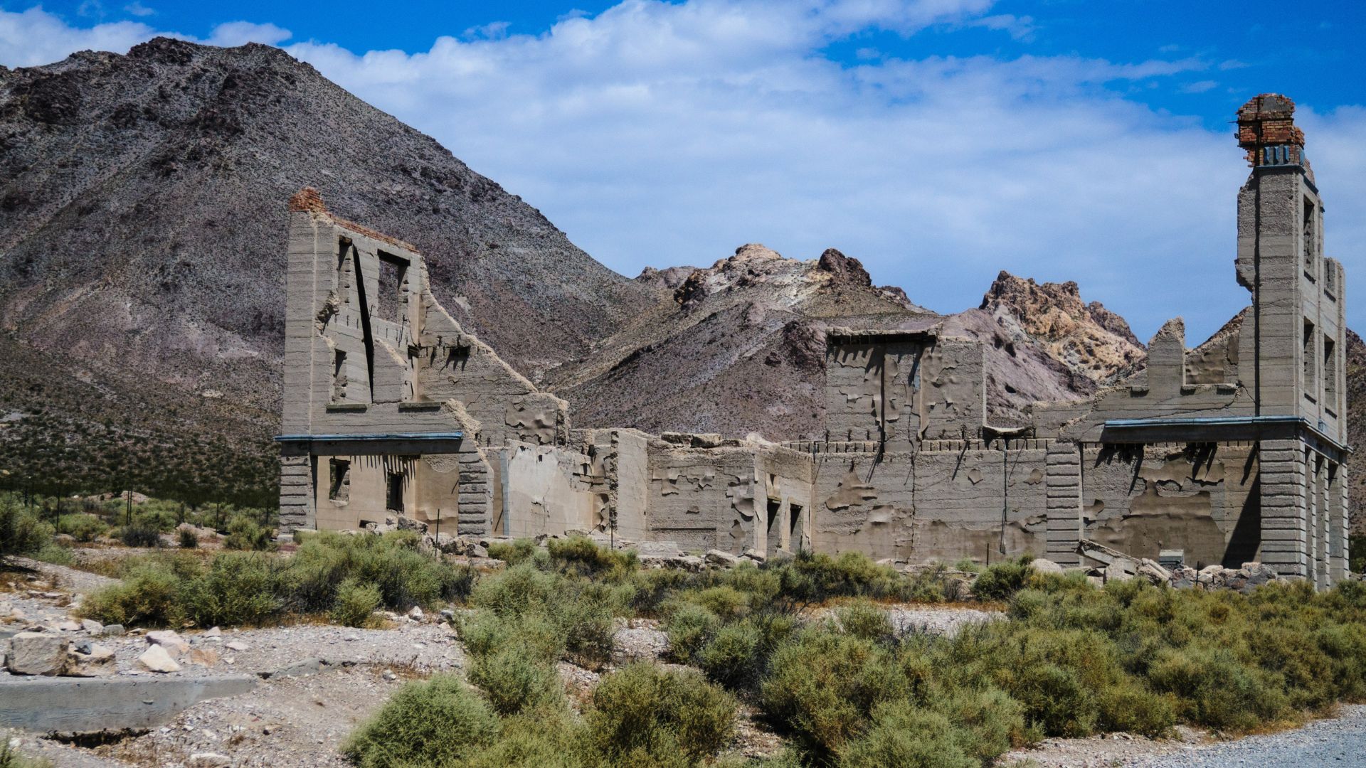 A wide shot captures the ruins of the Cook Bank Building and other structures in the Rhyolite Ghost Town in Nevada, with mountains rising behind them under a partially cloudy sky. The foreground features desert brush.