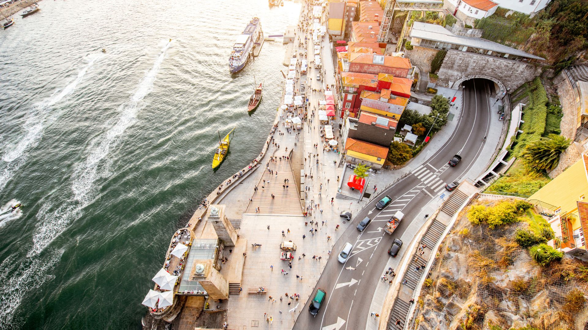 Aerial view of Porto's historic Ribeira district and the Douro River, showcasing colorful buildings, a bustling waterfront promenade, and a riverside road with a tunnel.