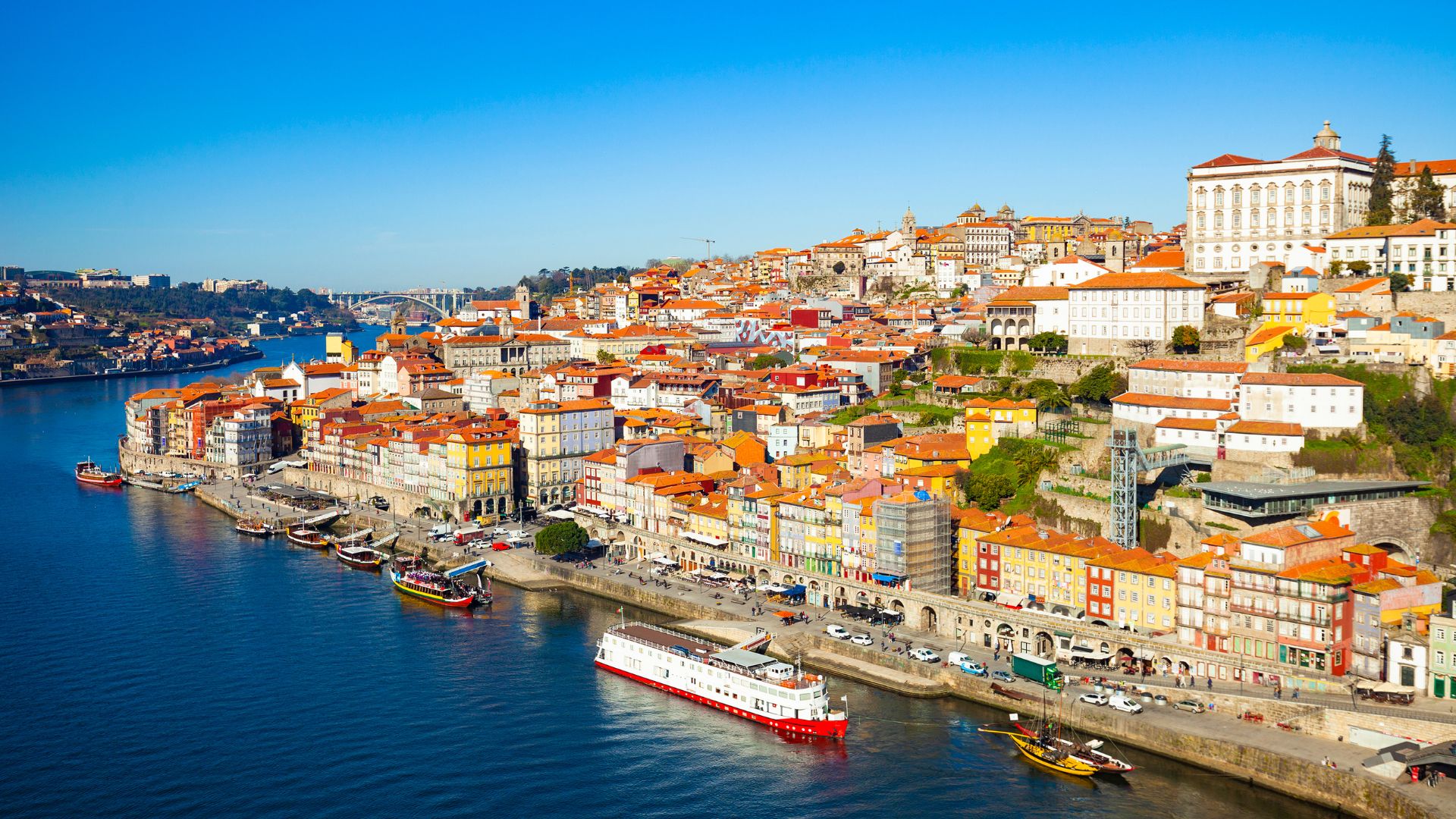 Panoramic view of Porto, Portugal, featuring the Douro River and colorful buildings. Traditional Rabelo boats are visible on the river. The city's historic architecture and vibrant atmosphere are captured in this scenic image.