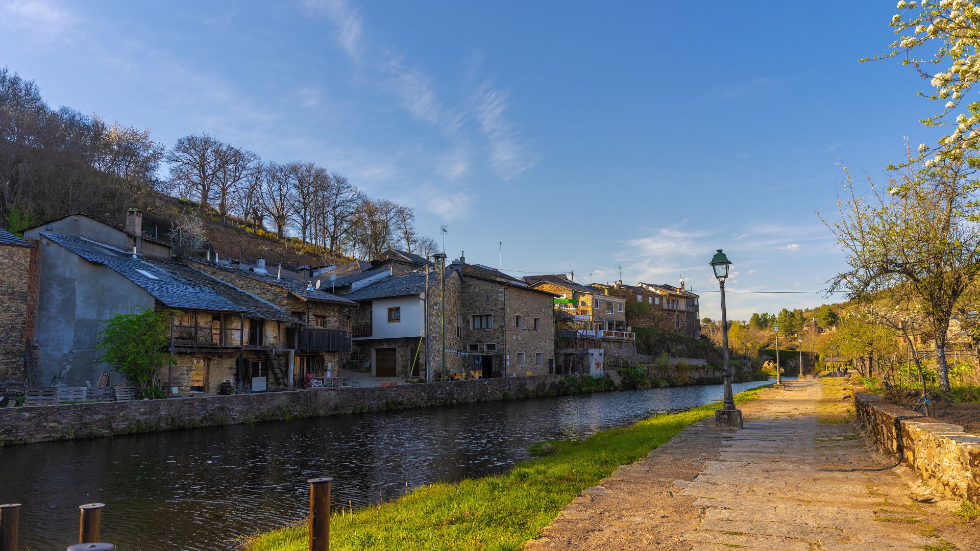 A picturesque view of the traditional village of Rio de Onor in Portugal, featuring stone houses with slate roofs lining the banks of the Onor River, with a clear blue sky overhead and lush green vegetation along the river path. 