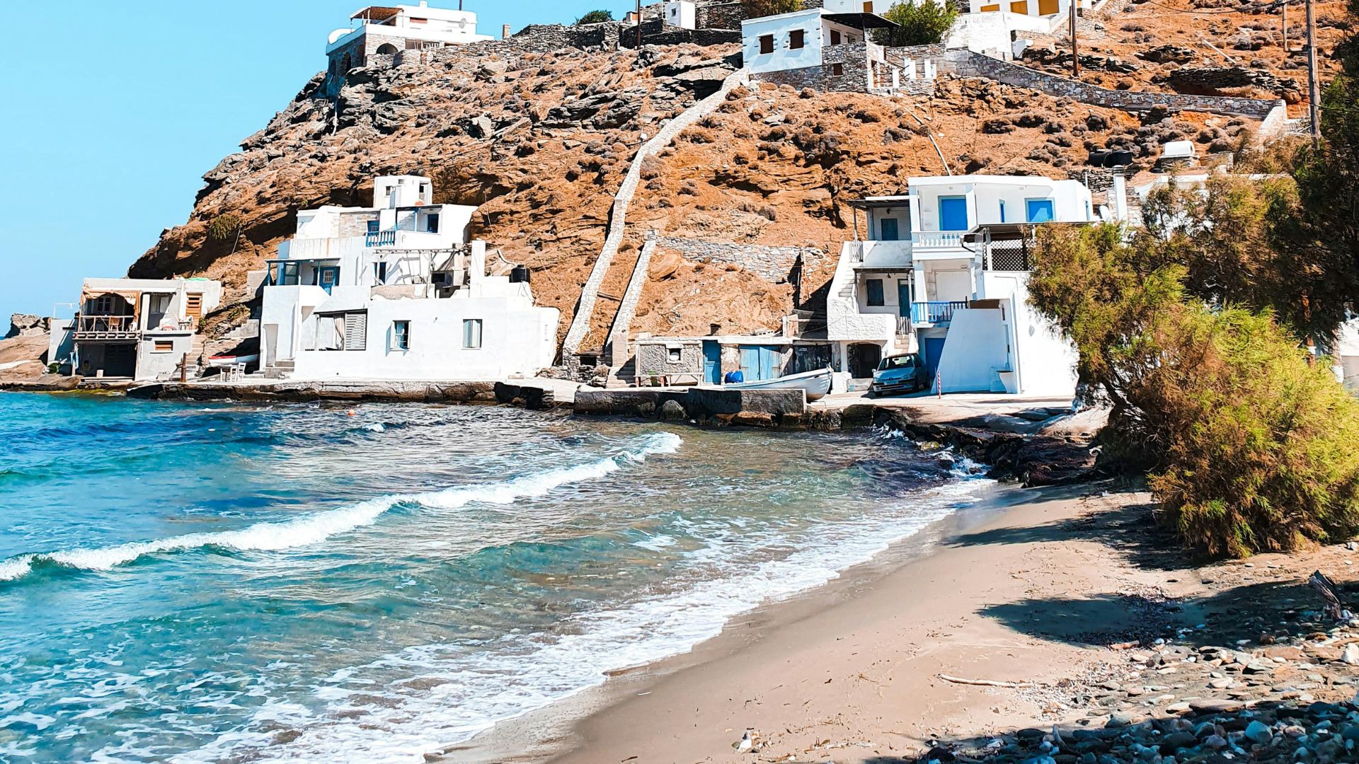 Rochari Beach in Tinos, Greece, with white buildings on a hill overlooking a sandy beach and the Aegean Sea.