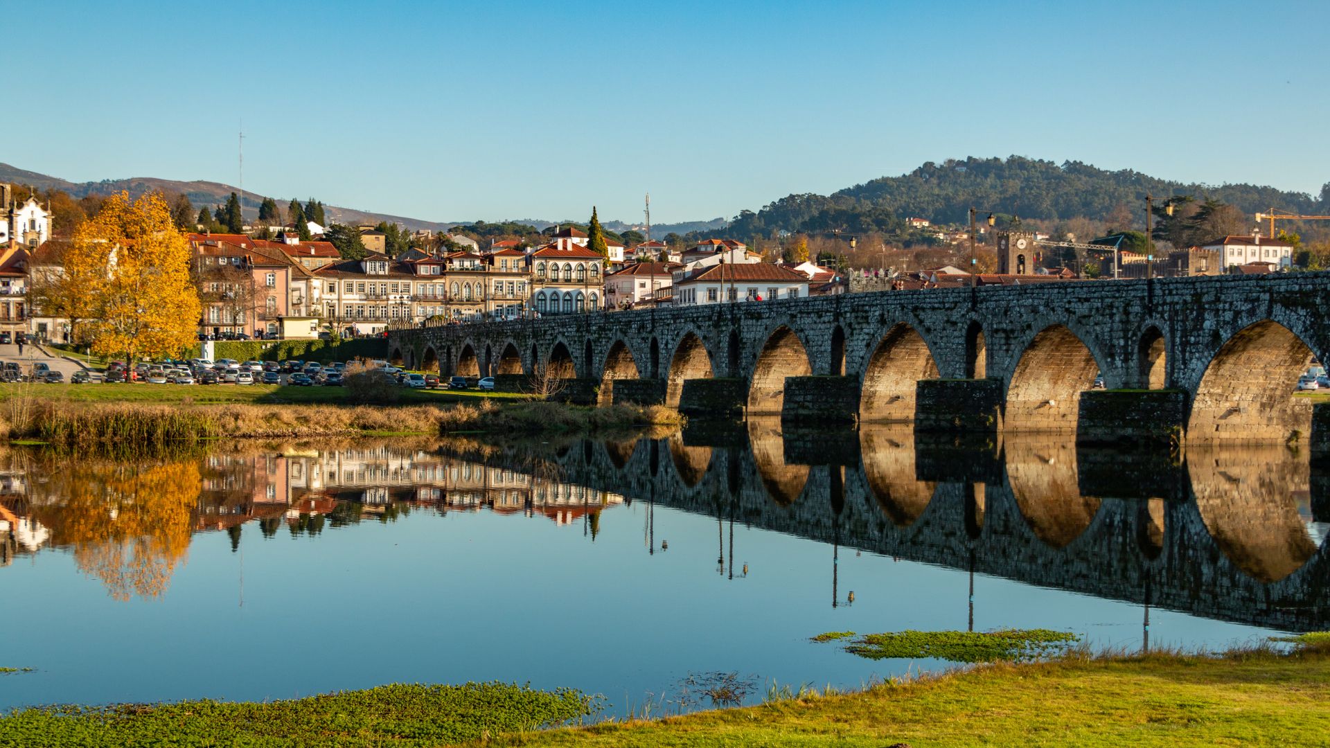 A panoramic view of the historic Roman and Medieval Bridge spanning the calm Lima River in Ponte de Lima, Portugal, with the town's charming buildings and autumn-colored trees reflected in the water under a clear blue sky.