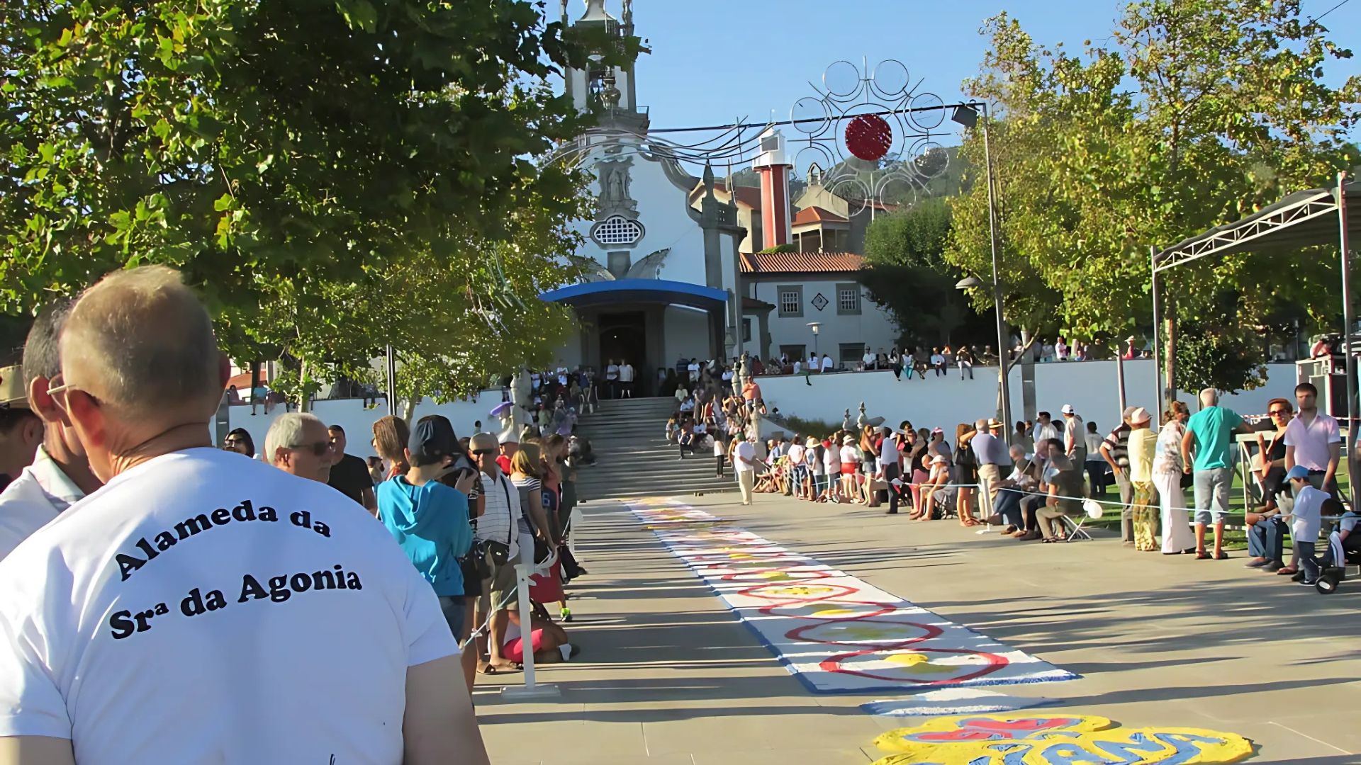  A crowd gathers in front of a church during the Romaria da Senhora d'Agonia festival in Viana do Castelo, with a decorated carpet on the ground and a man in the foreground wearing a shirt referencing "Alameda da Sra da Agonia."