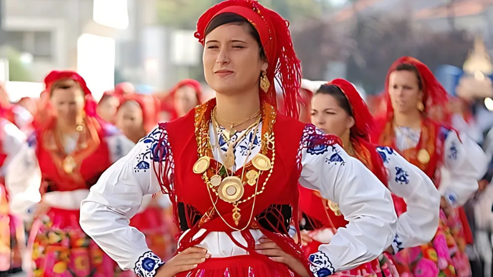A woman in a vibrant red and embroidered traditional Portuguese folk costume with elaborate gold jewelry and a red headscarf, standing on a cobblestone street with other similarly dressed individuals in the background.