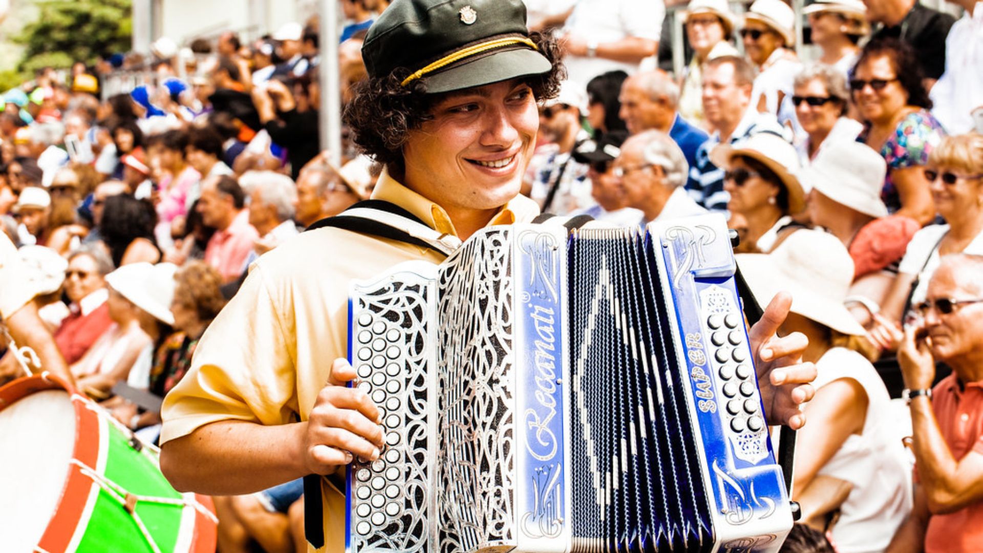 A young man in a yellow shirt and cap smiling while playing an accordion during a festival, with a crowd of people in the background.