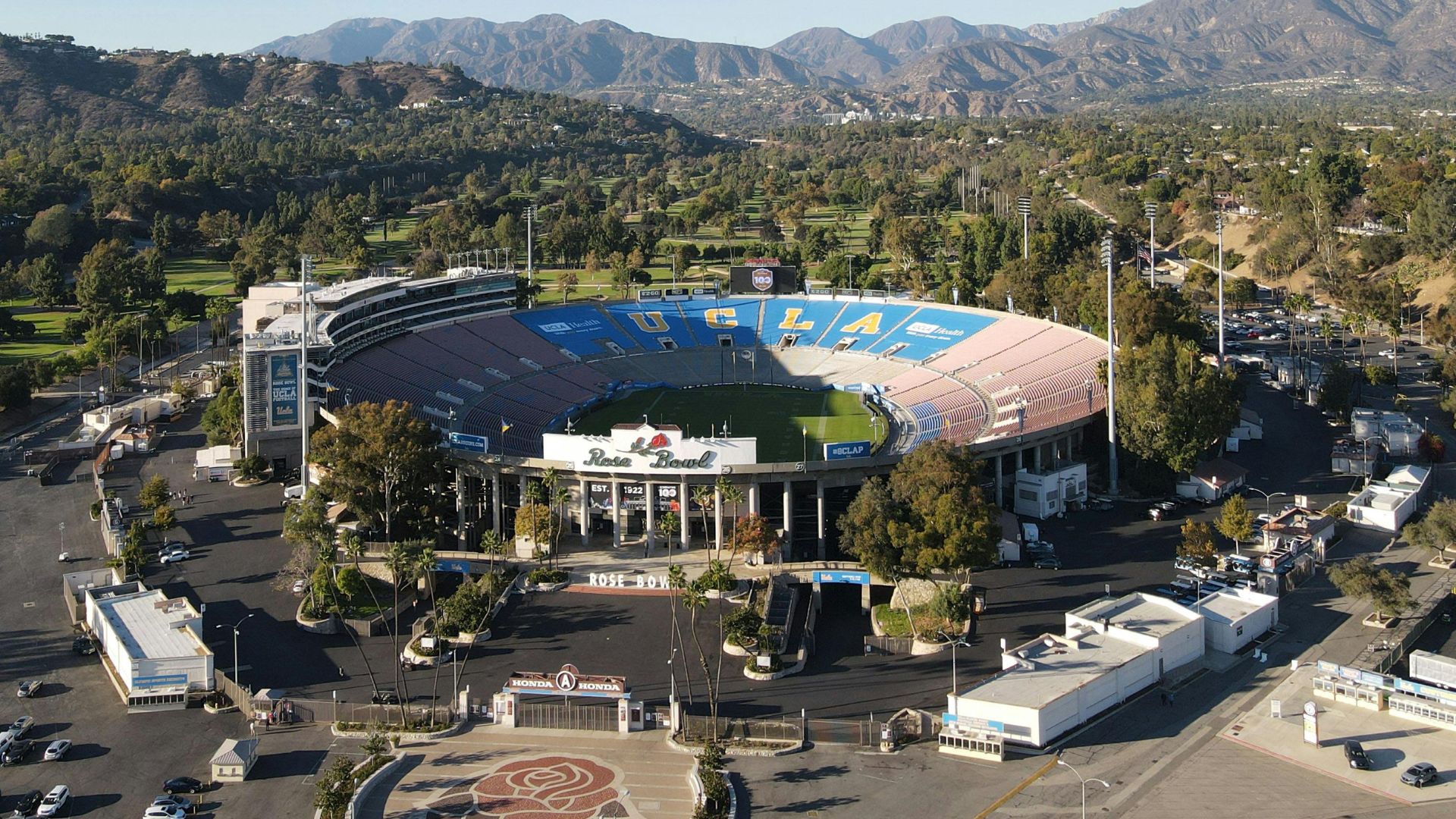 An aerial view of the Rose Bowl Stadium in Pasadena, California, surrounded by trees and with mountains in the background.