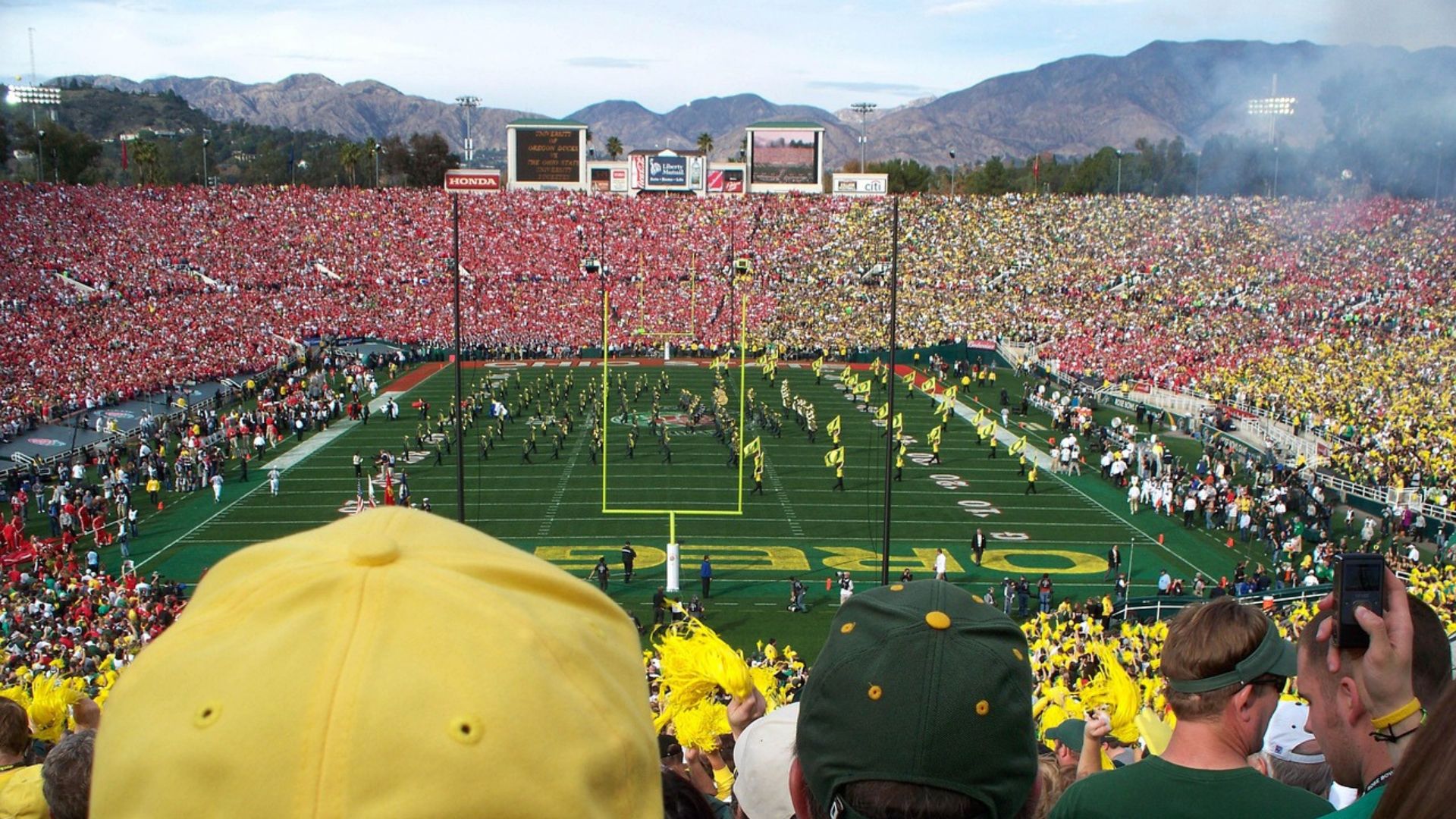 An aerial view of the Rose Bowl Stadium during a football game, with a large crowd in the stands and a marching band on the field.