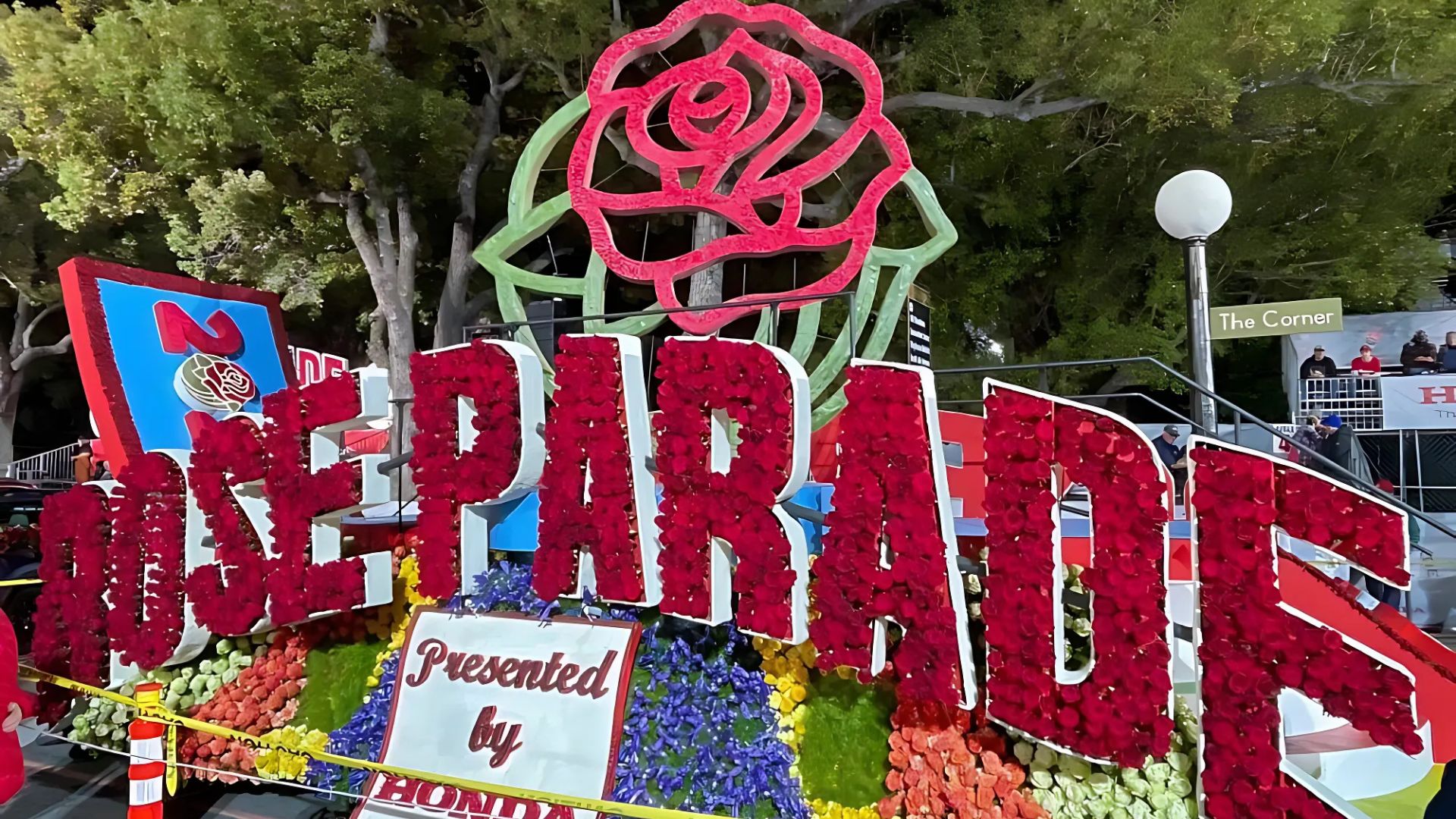 A large, colorful Rose Parade float with the words "ROSE PARADE Presented by HONDA" adorned with red roses and other flowers, set against a backdrop of trees and a night sky. A large red rose graphic is above the text, and "The Corner" sign is visible in the background on the right.