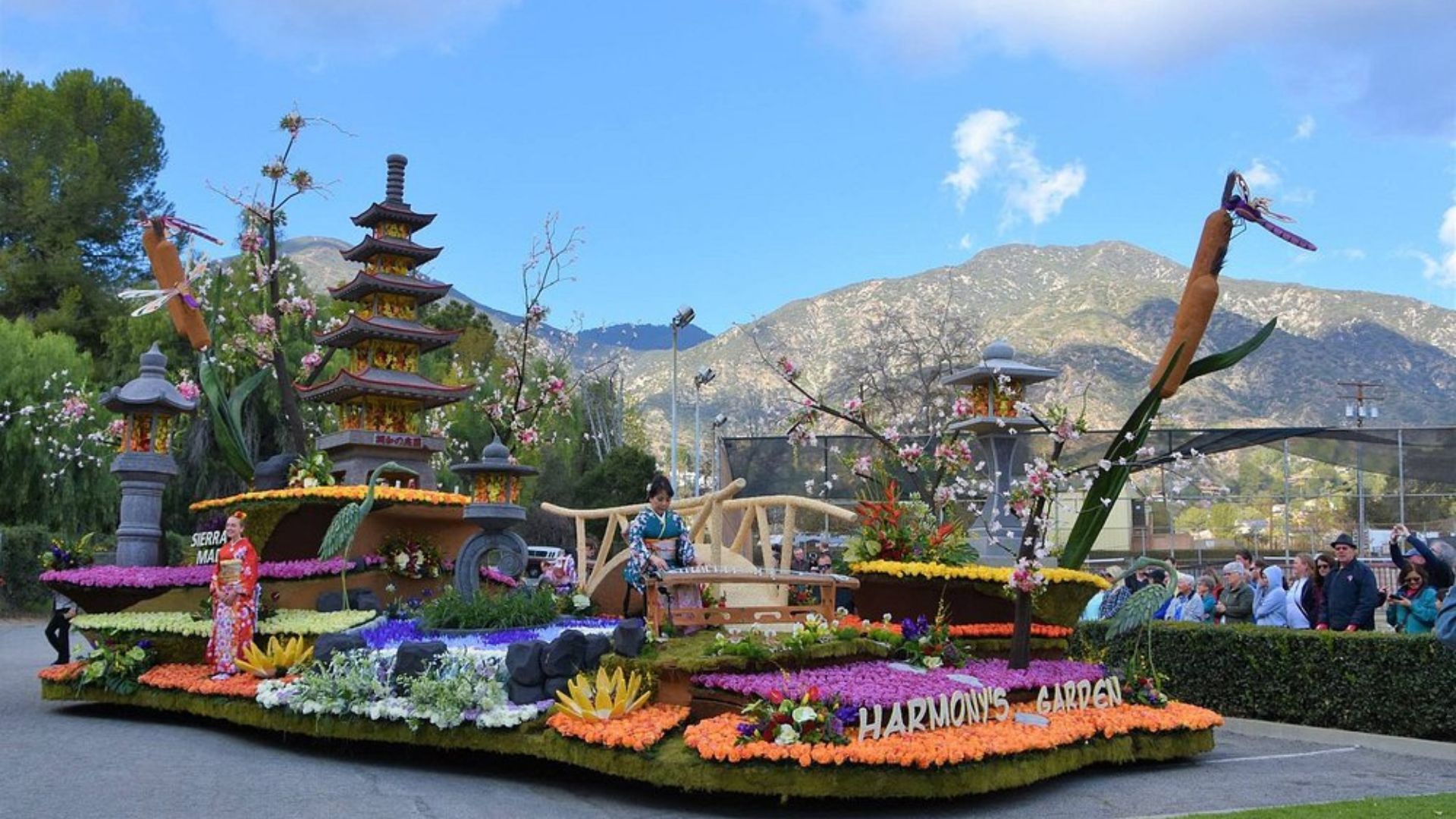 A colorful Rose Parade float featuring a Japanese-style pagoda, a bridge, and a garden scene with various flowers and foliage, set against a backdrop of mountains and a blue sky.