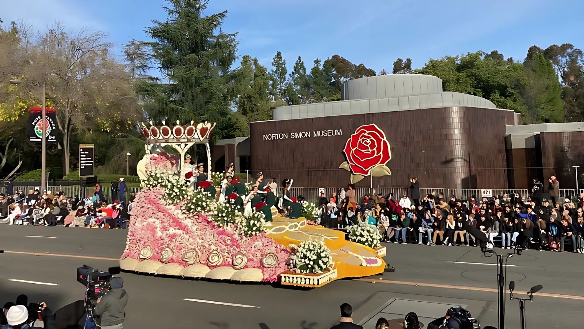 A large, ornate float decorated with flowers, resembling a crown and a shoe, moves down a street during the Rose Parade in Pasadena, California, with the Norton Simon Museum building visible in the background.