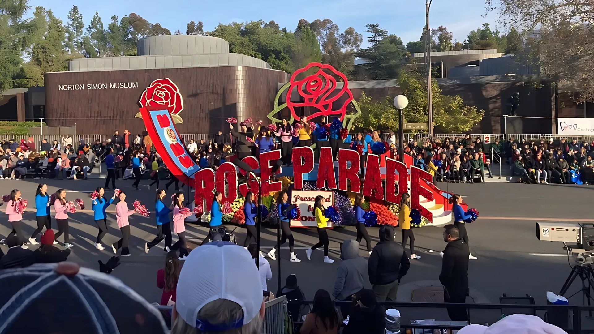 A vibrant Rose Parade float adorned with flowers and featuring the words "ROSE PARADE" passes by a crowd, with the Norton Simon Museum visible in the background.