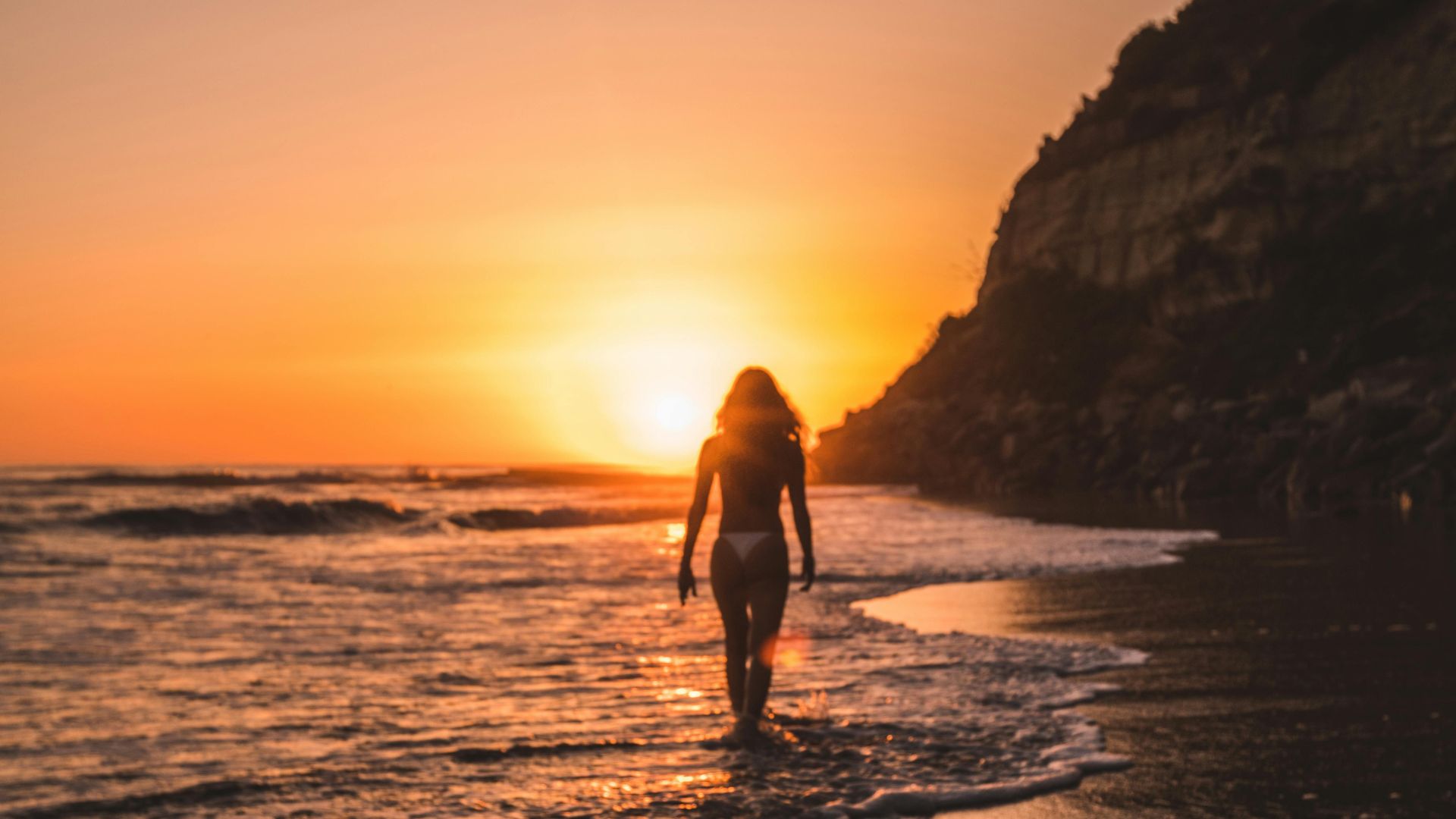 Woman in a bikini walking on a San Diego beach at sunset, silhouetted against the vibrant orange sky and ocean waves, with a cliffside in the background.