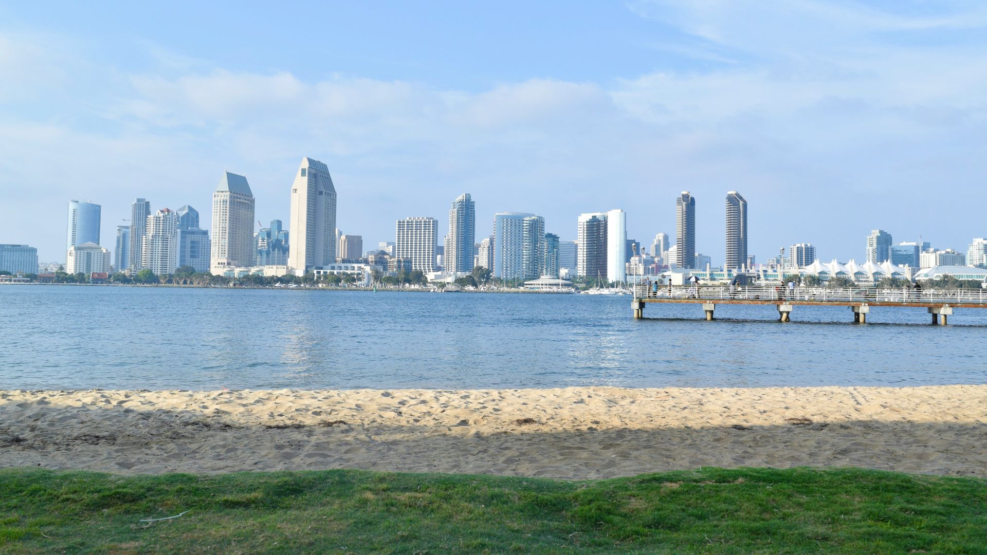 San Diego skyline from Coronado Island beach, featuring downtown skyscrapers, a calm bay, and a sandy shore with green grass in the foreground.