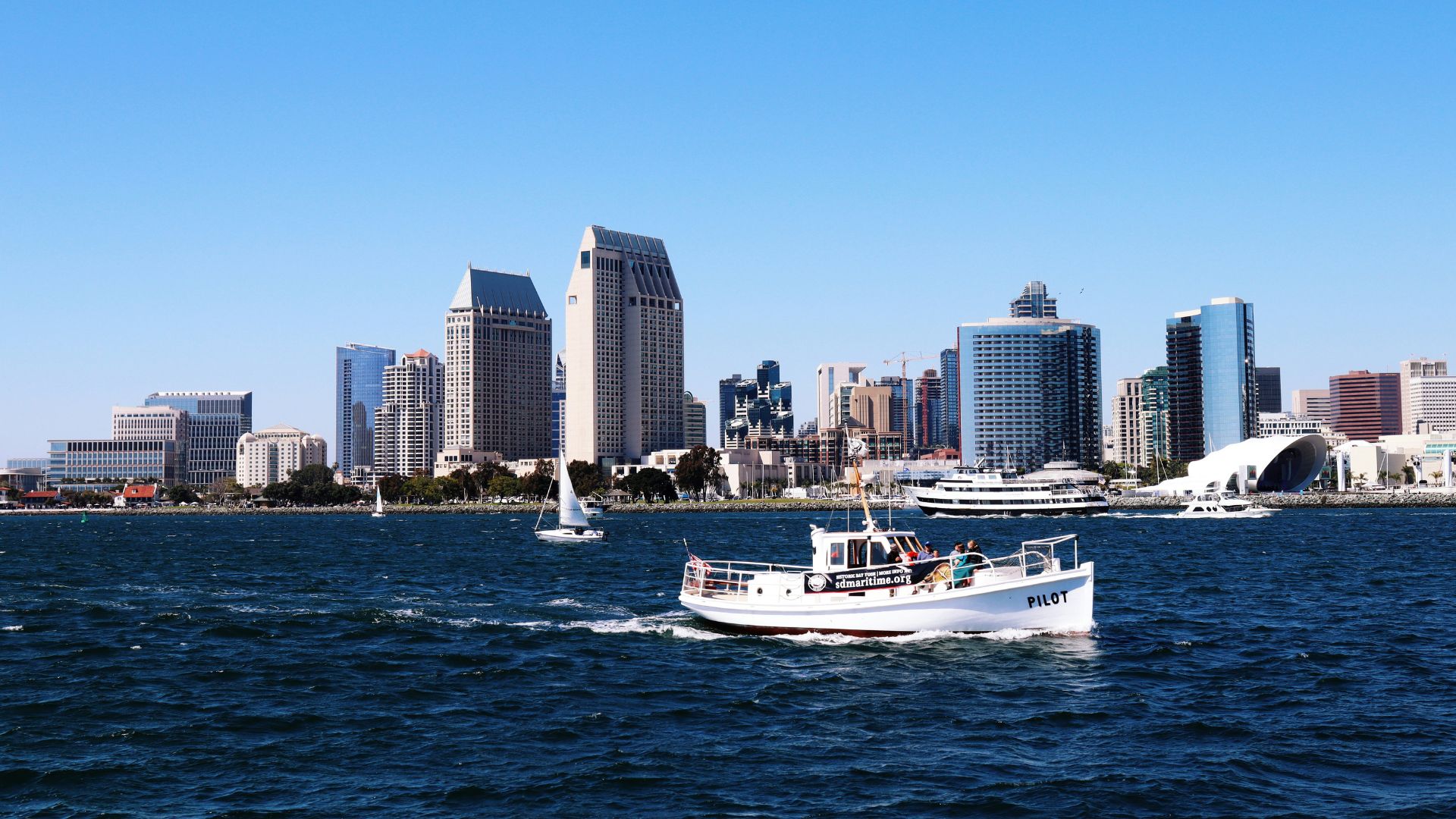 A white pilot boat navigates the waters of San Diego Bay with the downtown San Diego skyline visible in the background under a clear blue sky.