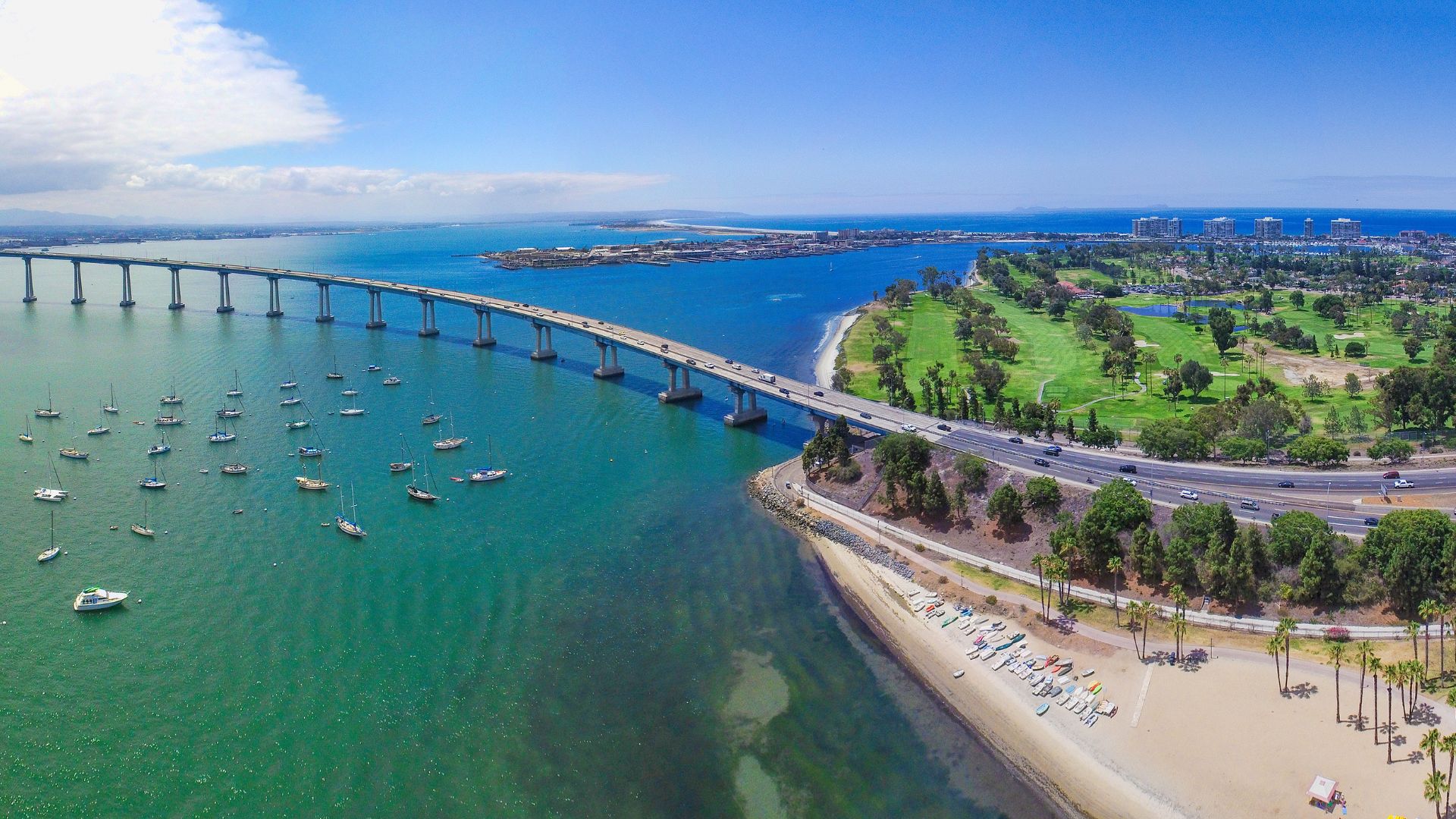 An aerial view of the San Diego–Coronado Bridge spanning across a bay, with a golf course and beachfront visible on the right, and numerous boats anchored in the water on the left.