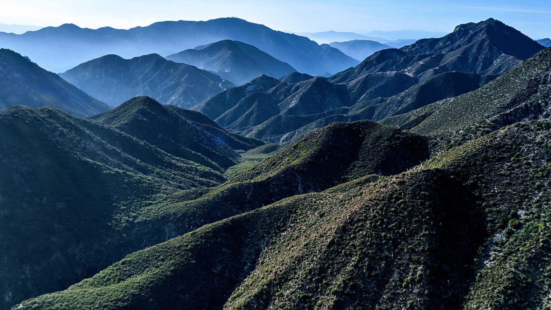 Scenic view of the San Gabriel Mountains within the Angeles National Forest in Southern California. The image showcases layers of mountains with lush vegetation and a hazy blue sky, highlighting the natural beauty and biodiversity of the region. Ideal for outdoor enthusiasts and nature lovers.