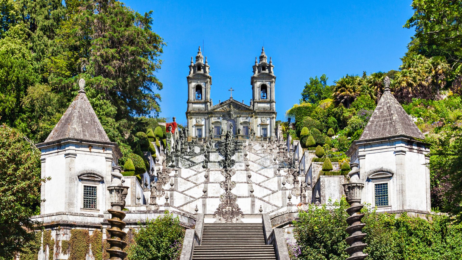 A grand Baroque staircase leading up a hill to a large church with twin bell towers, surrounded by lush green trees.