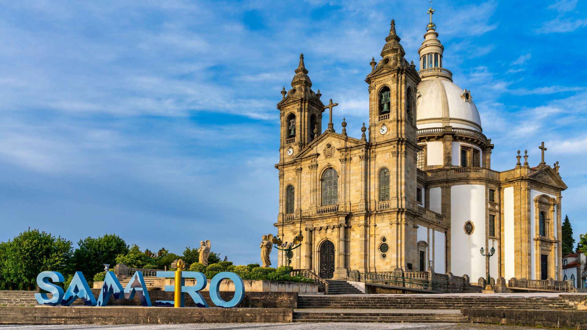 A large, ornate neo-Baroque basilica with a prominent dome and two bell towers, set against a blue sky with clouds, with a "SAMEIRO" sign in the foreground.