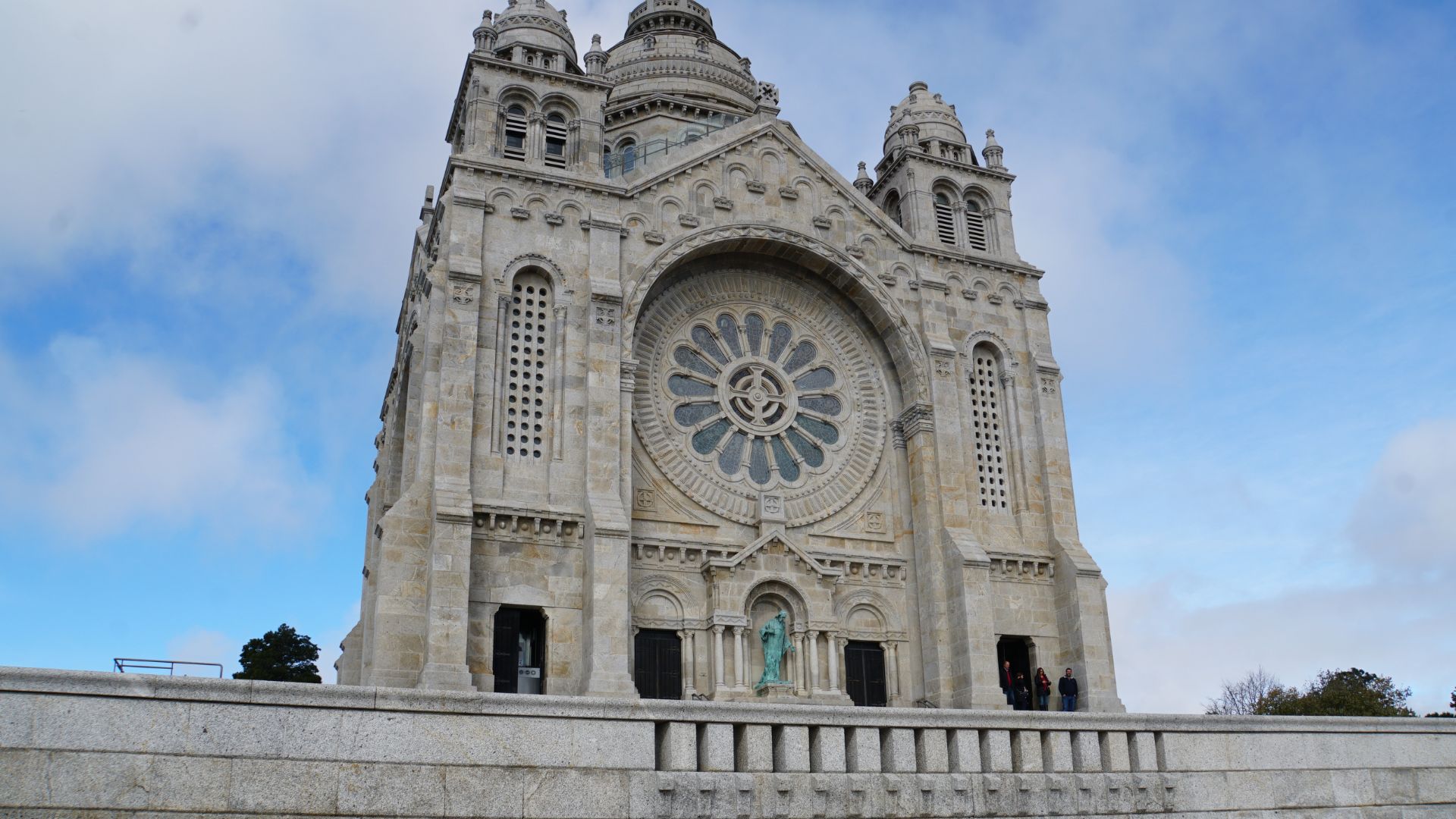 A grand stone sanctuary with a large rose window and imposing facade, set against a blue sky with clouds.