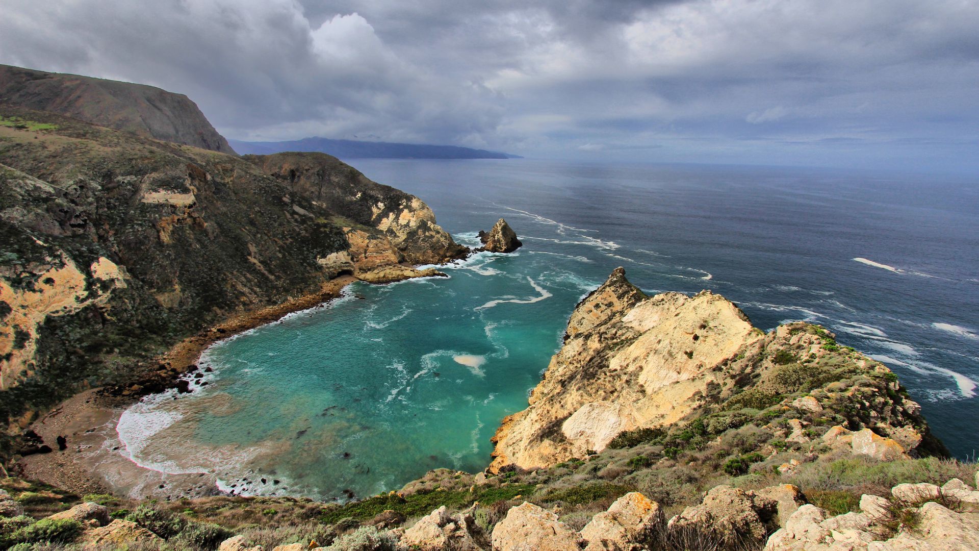 A high-angle view overlooking a turquoise cove with a sandy beach and rocky cliffs, leading out to a vast expanse of dark blue ocean under a cloudy, dramatic sky. In the foreground, rugged, scrub-covered terrain slopes down towards the cove.
