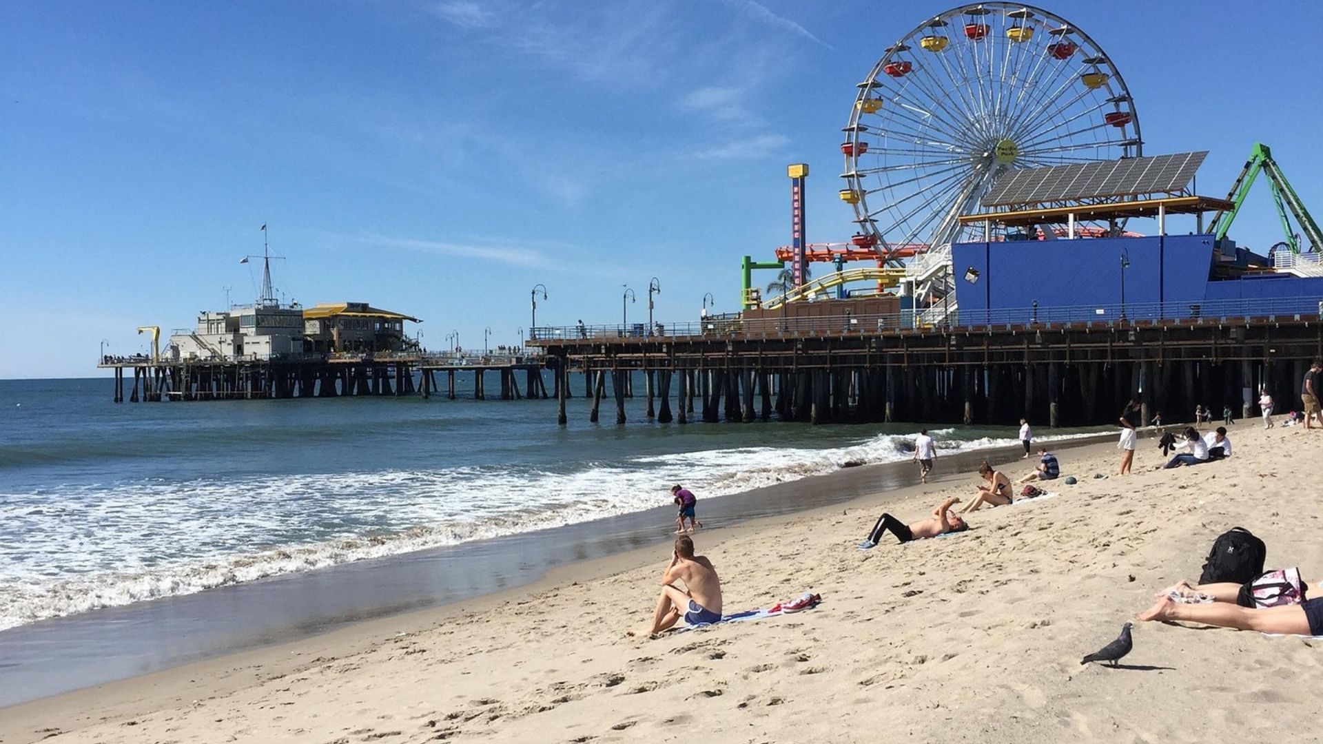 People laying down on the white sand of Santa Monica Beach in Los Angeles, Southern California with Santa Monica Pier and blue water in the background.