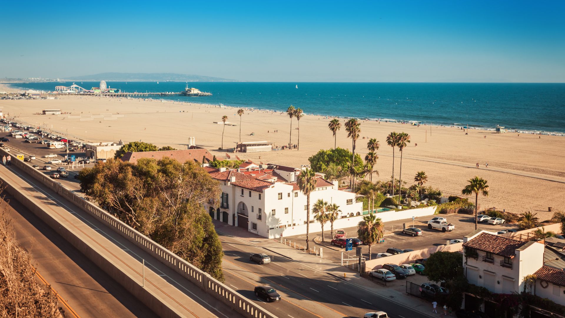 Aerial view of Santa Monica State Beach and Pier in California, featuring golden sand, the iconic pier with Pacific Park, the Pacific Ocean, and coastal buildings alongside a highway, under a clear blue sky.walk